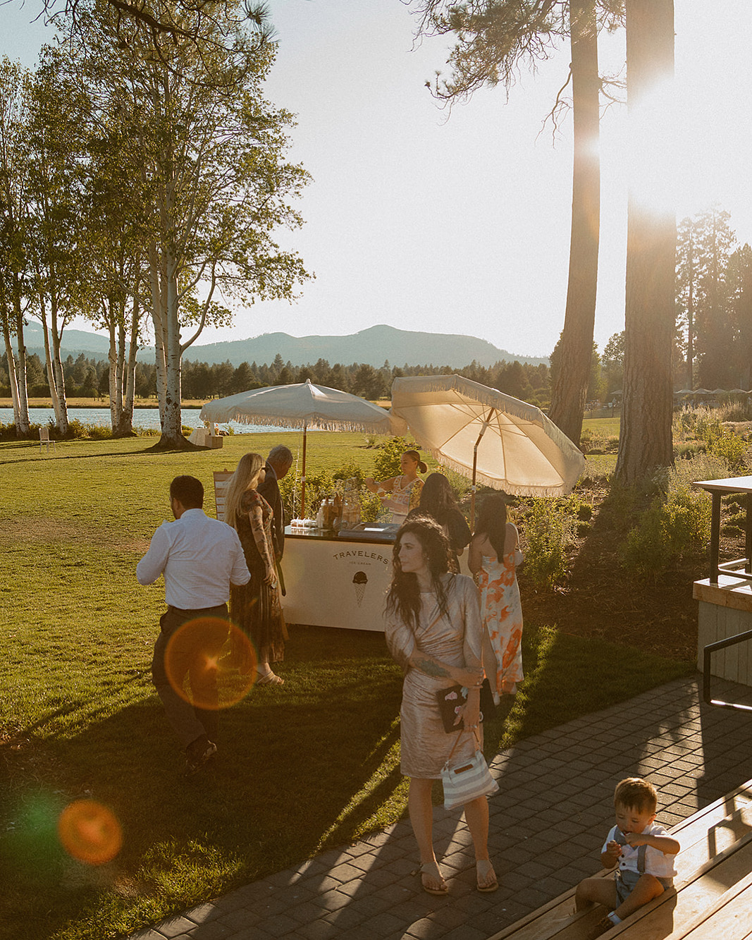People gather outdoors near a table with an umbrella, trees and sunlight in the background, and a child sitting on the ground.