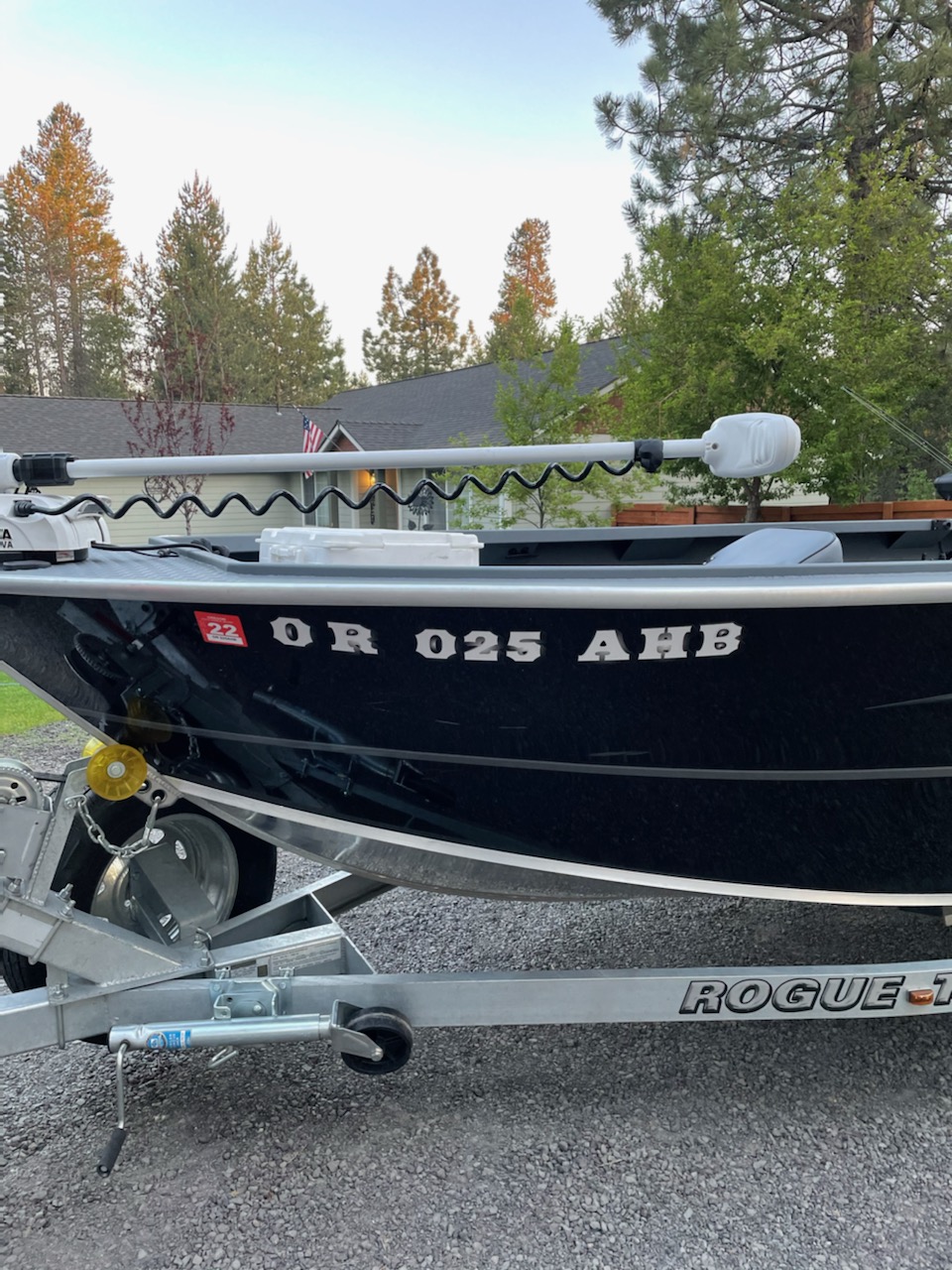 Boat on trailer with registration number, surrounded by trees and houses, under a clear sky.