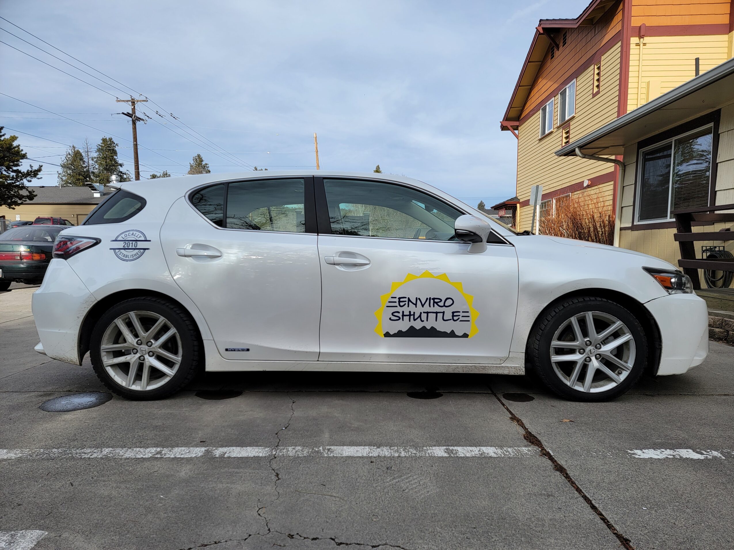 White sedan with a yellow and black logo on the door, parked in a lot with buildings and trees in background.
