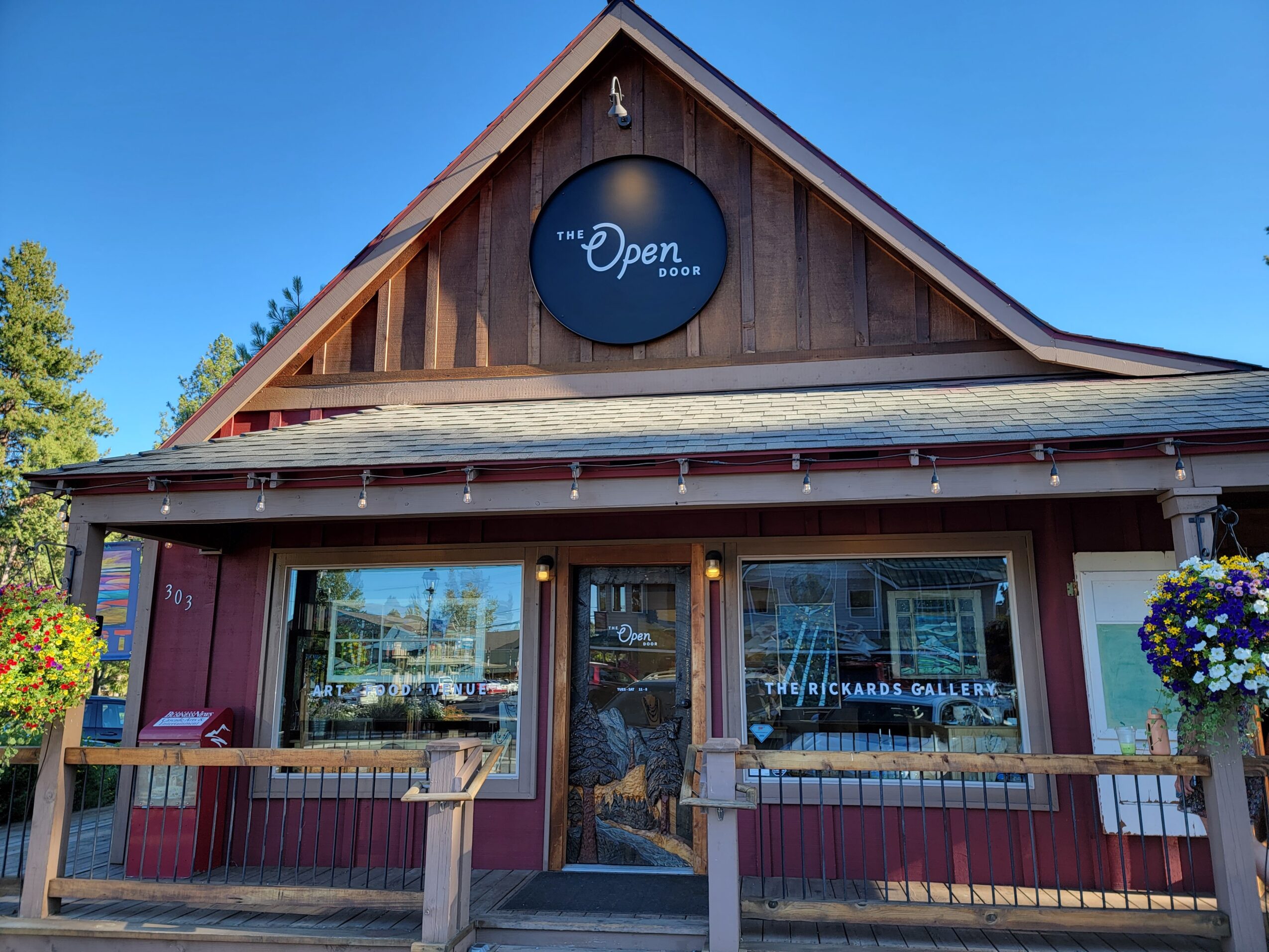 Front view of a building with a sign that says 'Open,' two large windows, and hanging flower baskets on each side.