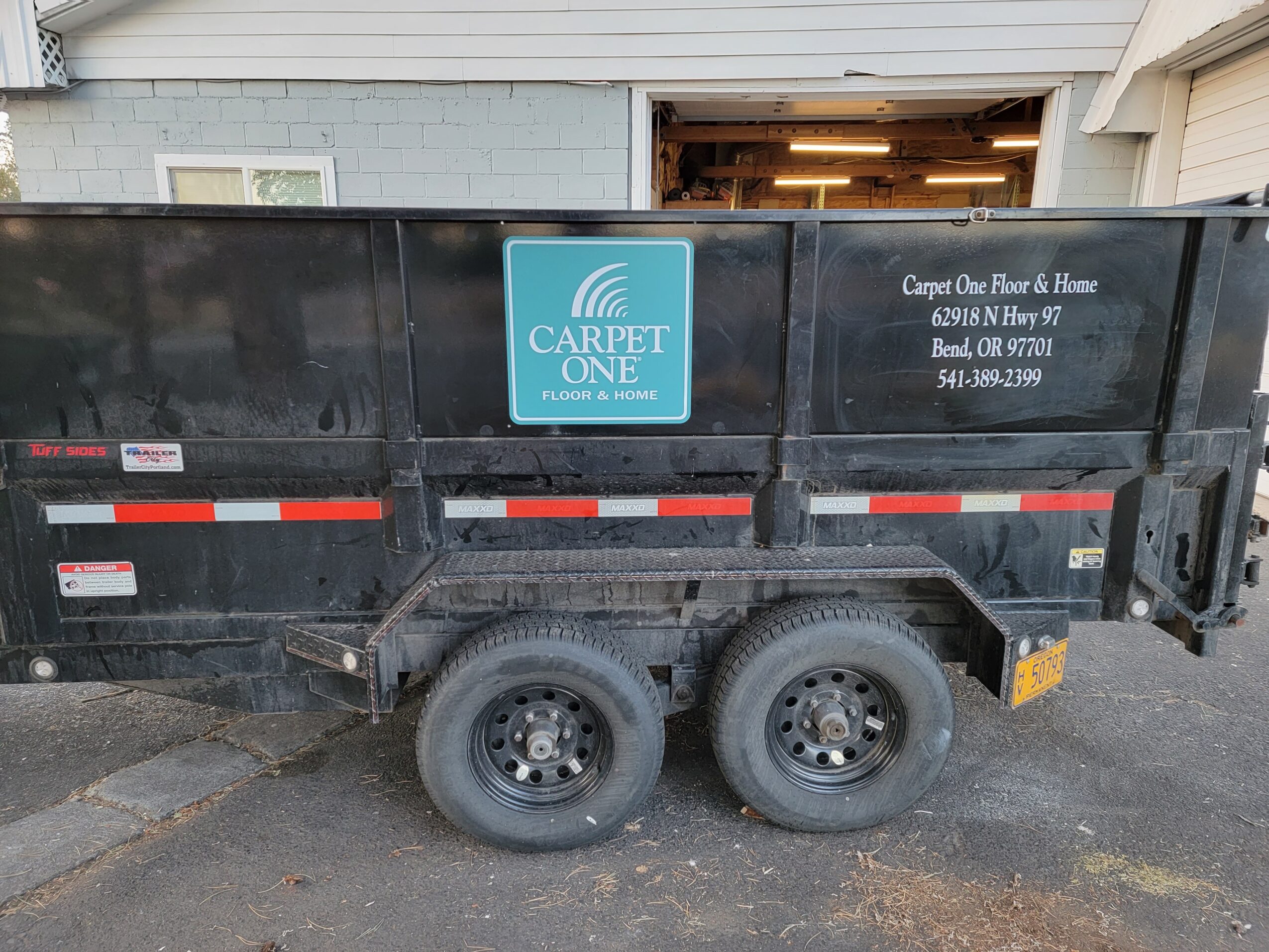 Black trailer with a blue Carpet One logo, parked outside a garage, with two wheels visible, on a paved surface.