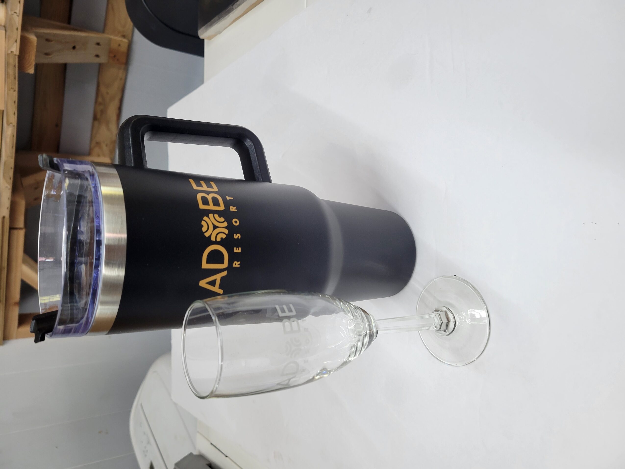A black thermos with gold text, a clear glass, and a wine glass on a white surface.