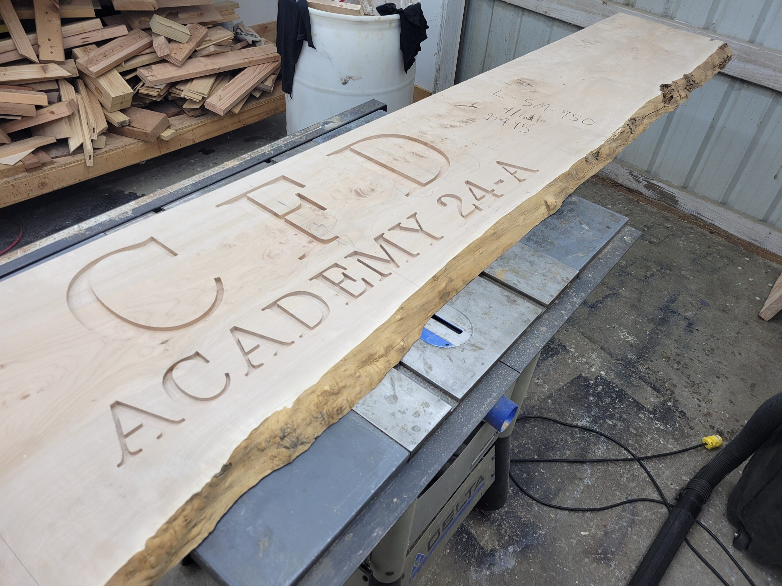 Wooden board with engraved text, leaning against a wall, with a pile of wood pieces nearby.