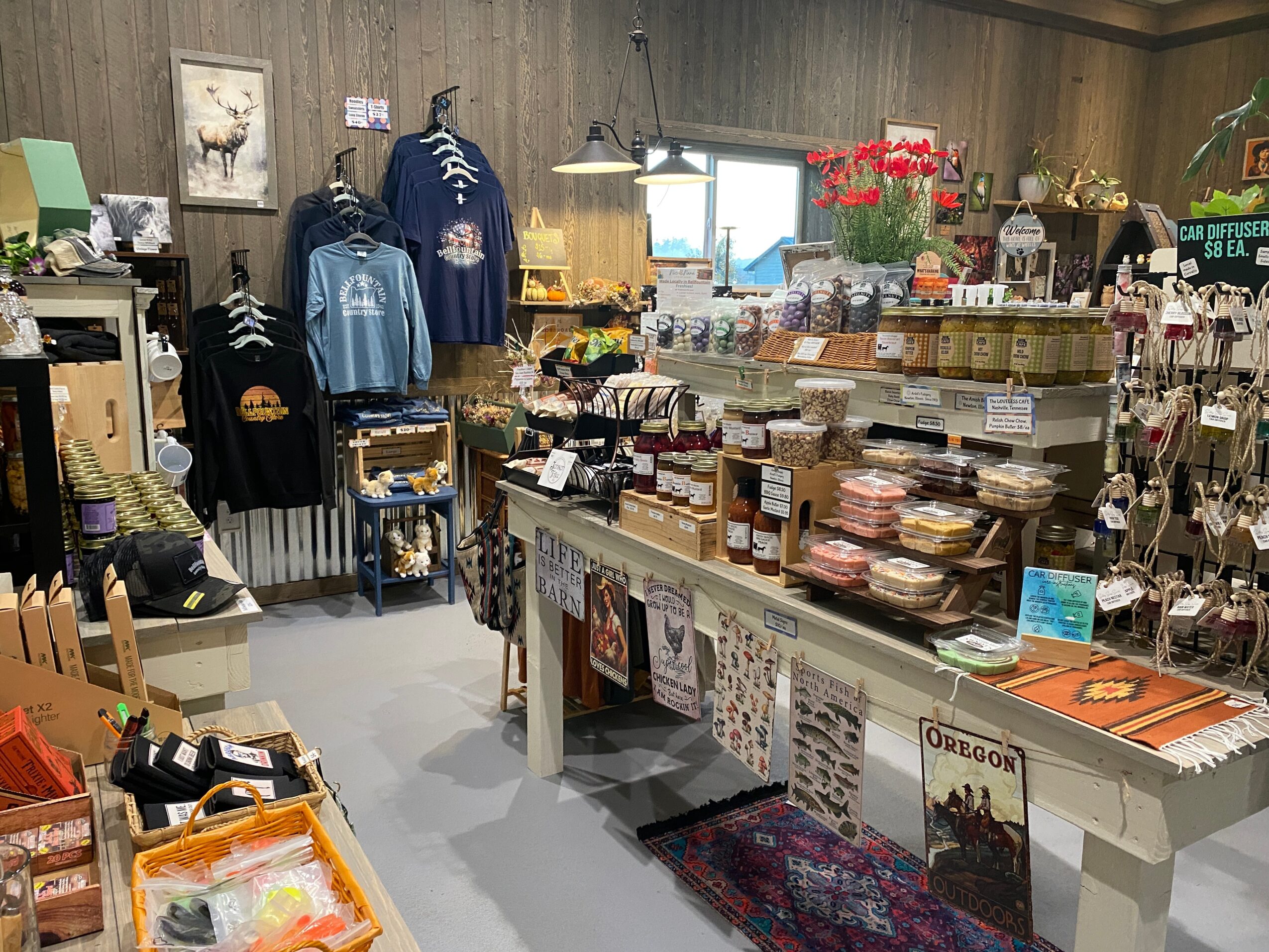 Indoor store with clothing, souvenirs, and gift items on shelves and tables, wooden walls, and a window with flowers.