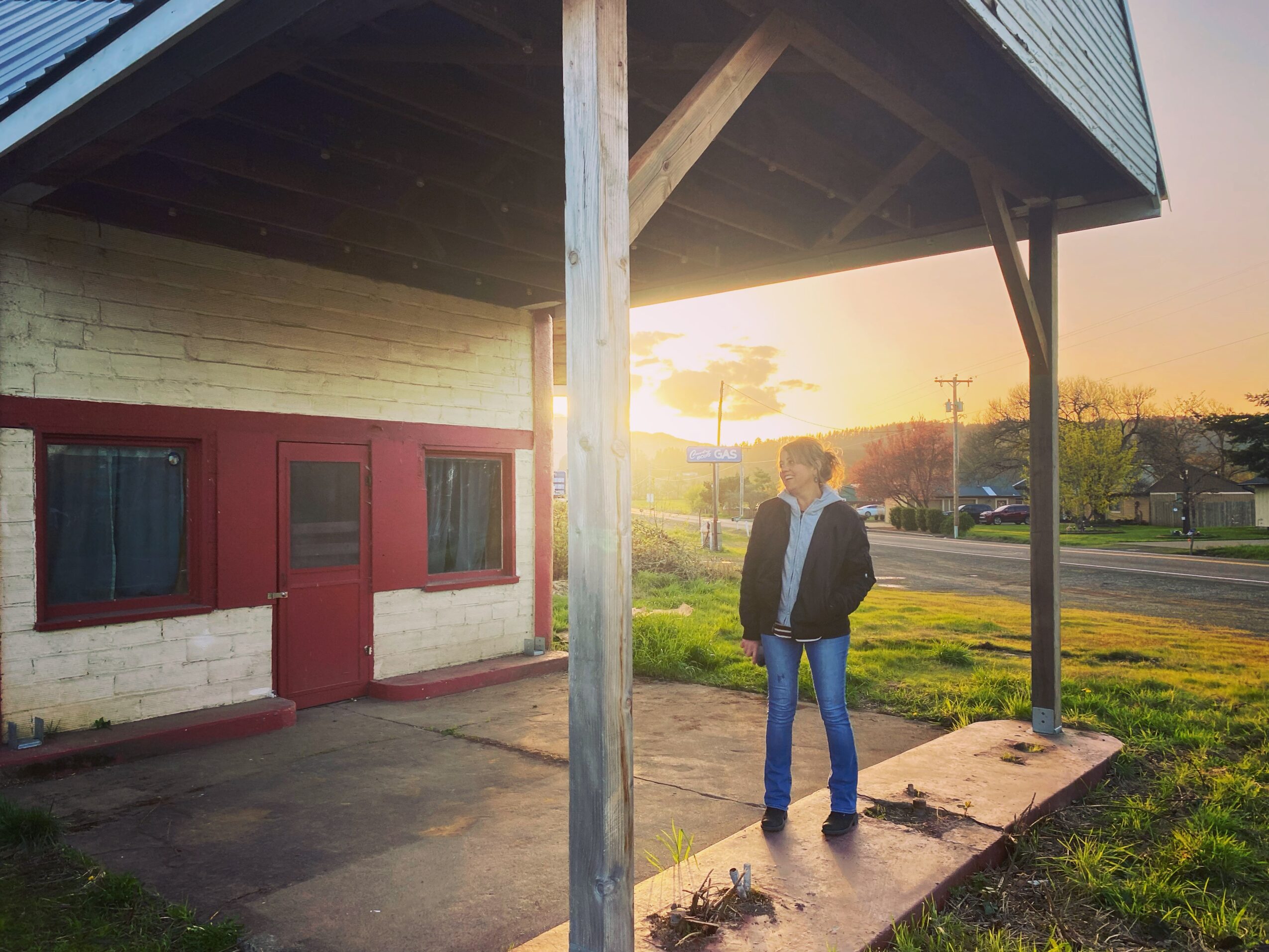 Person standing outside a building with a red door, sunlight in the background, grassy area nearby.