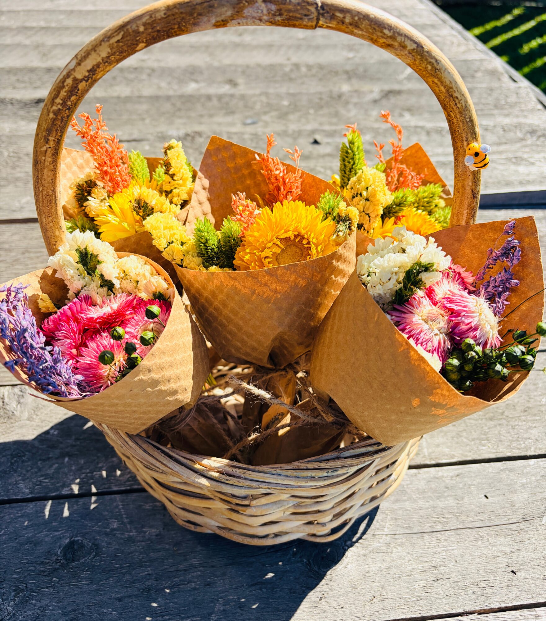 Colorful flower bouquets in a wicker basket on a wooden surface, with a handle and sunlight shining.
