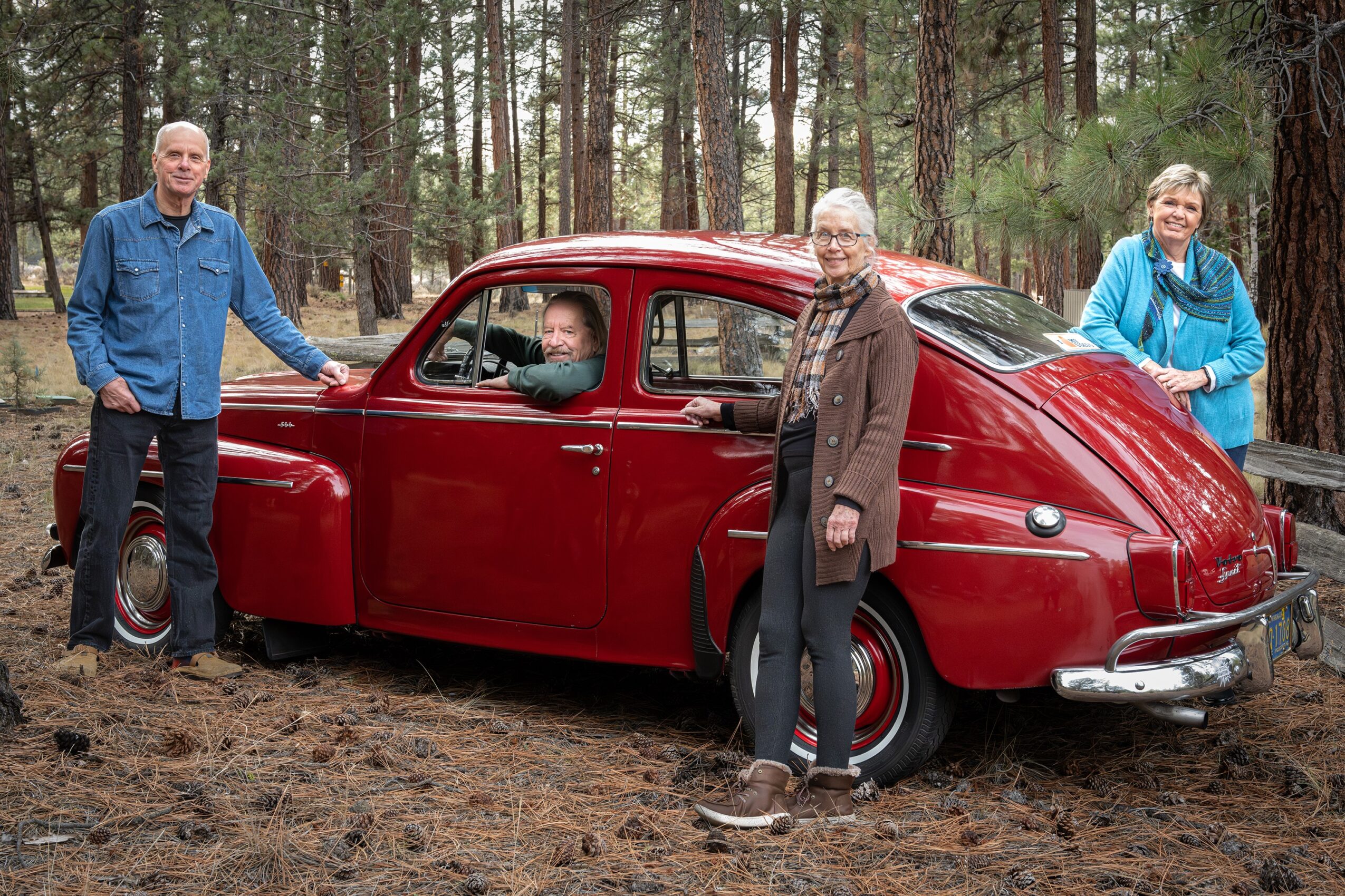 Four people and a vintage red car in a forest, with trees and ground visible.