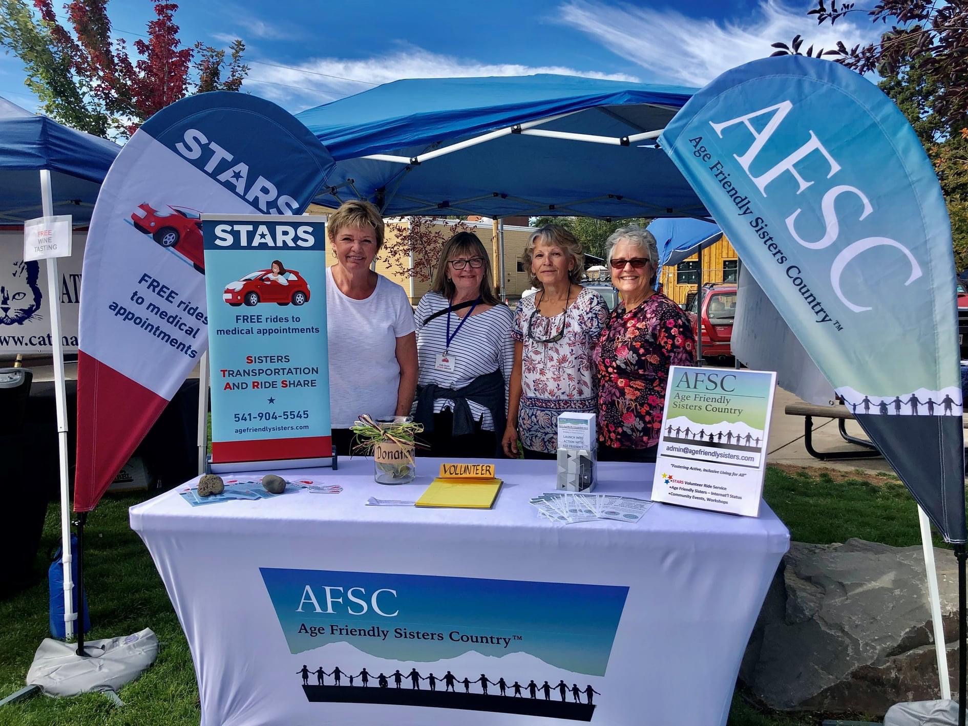 Four women stand behind an AFSC booth with banners, under a blue canopy, outdoors on a sunny day.