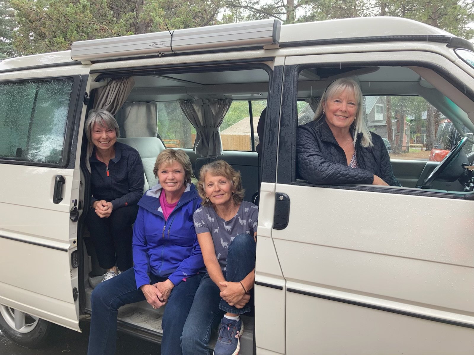 Four women sitting and standing in and outside a white camper van, smiling outdoors with trees in the background.