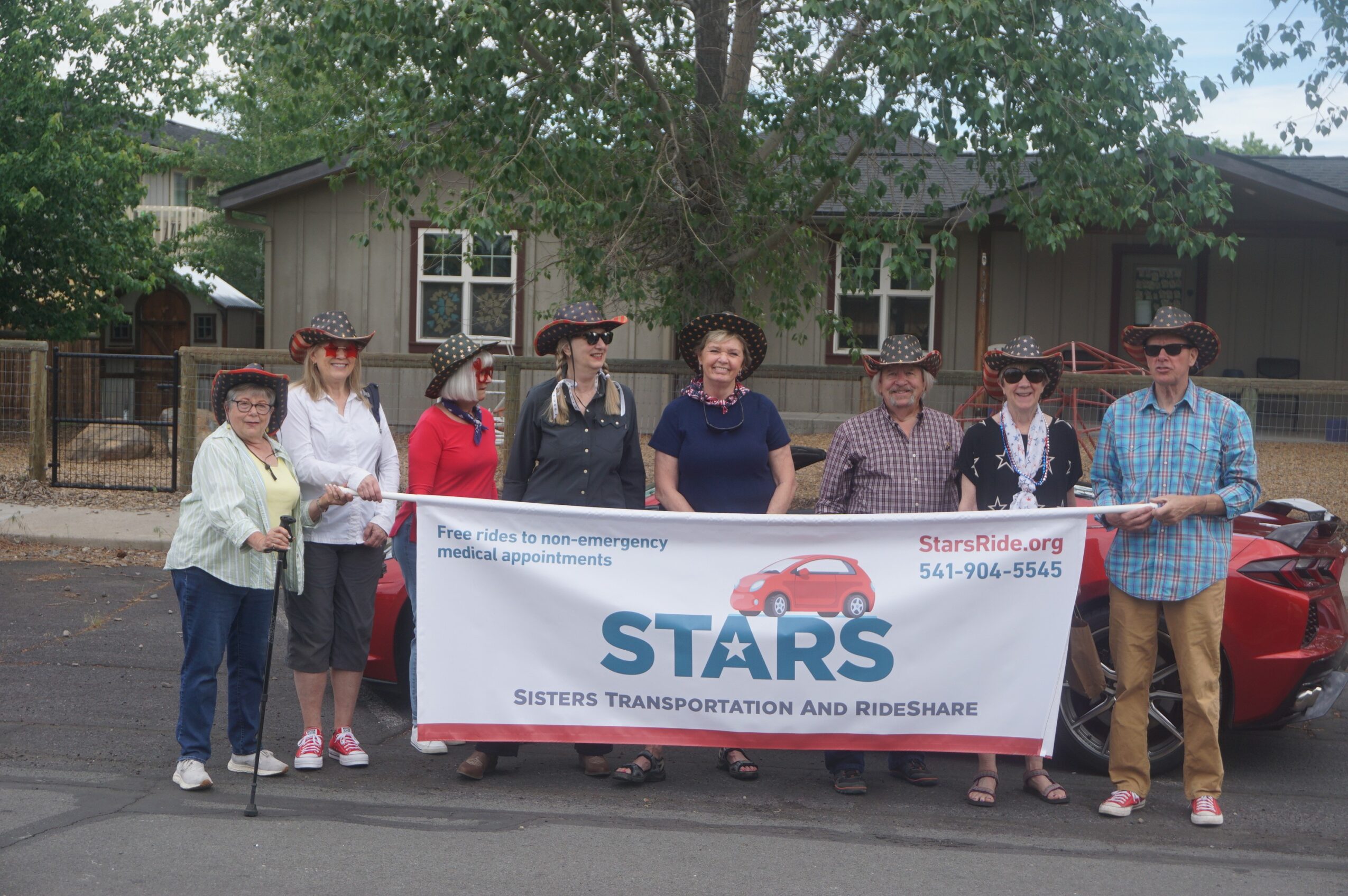 Group of eight people holding a banner in front of a house and trees, participating in a transportation event.