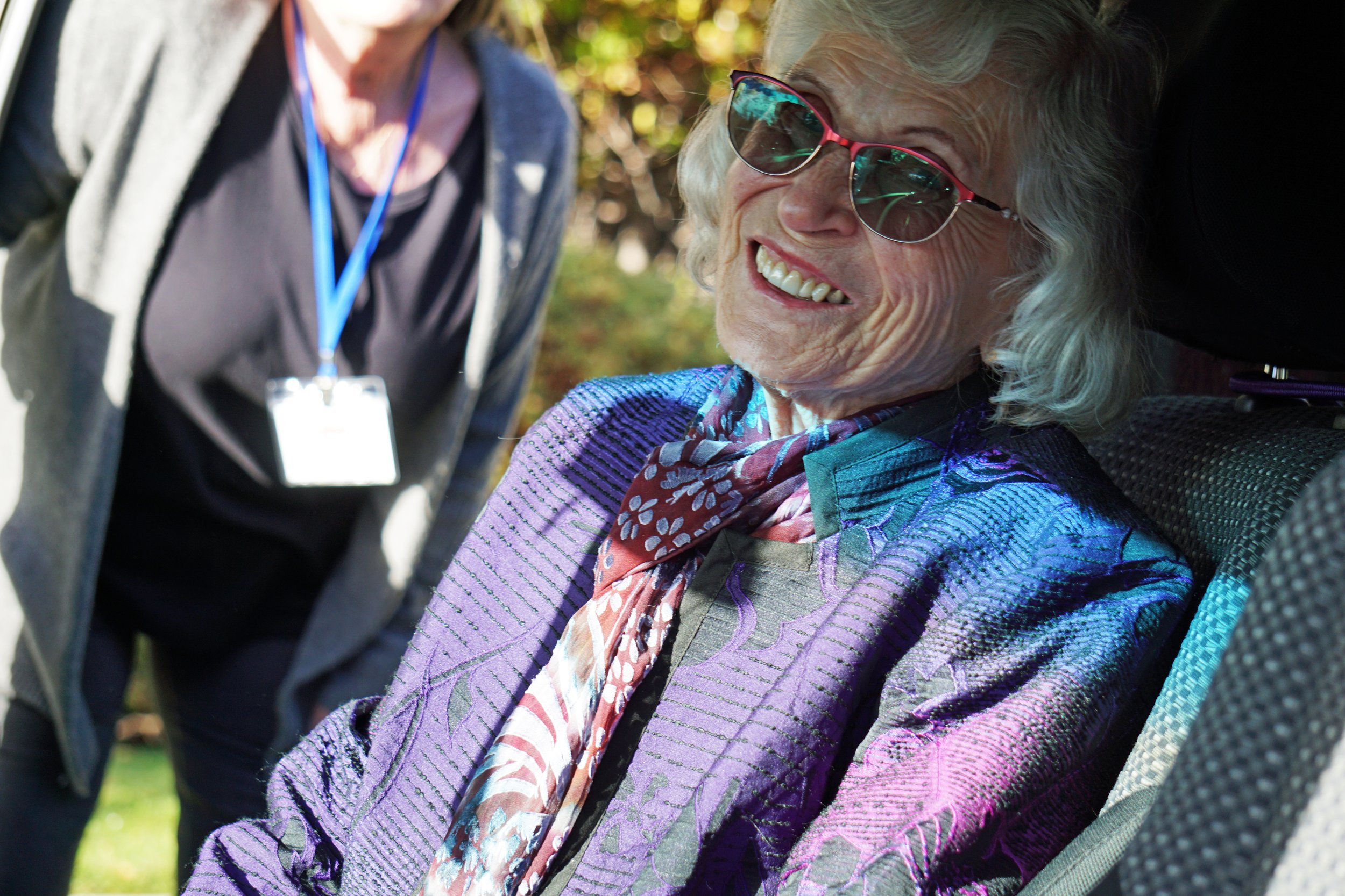 Smiling elderly woman with glasses and curly hair outdoors, wearing a purple jacket and scarf.