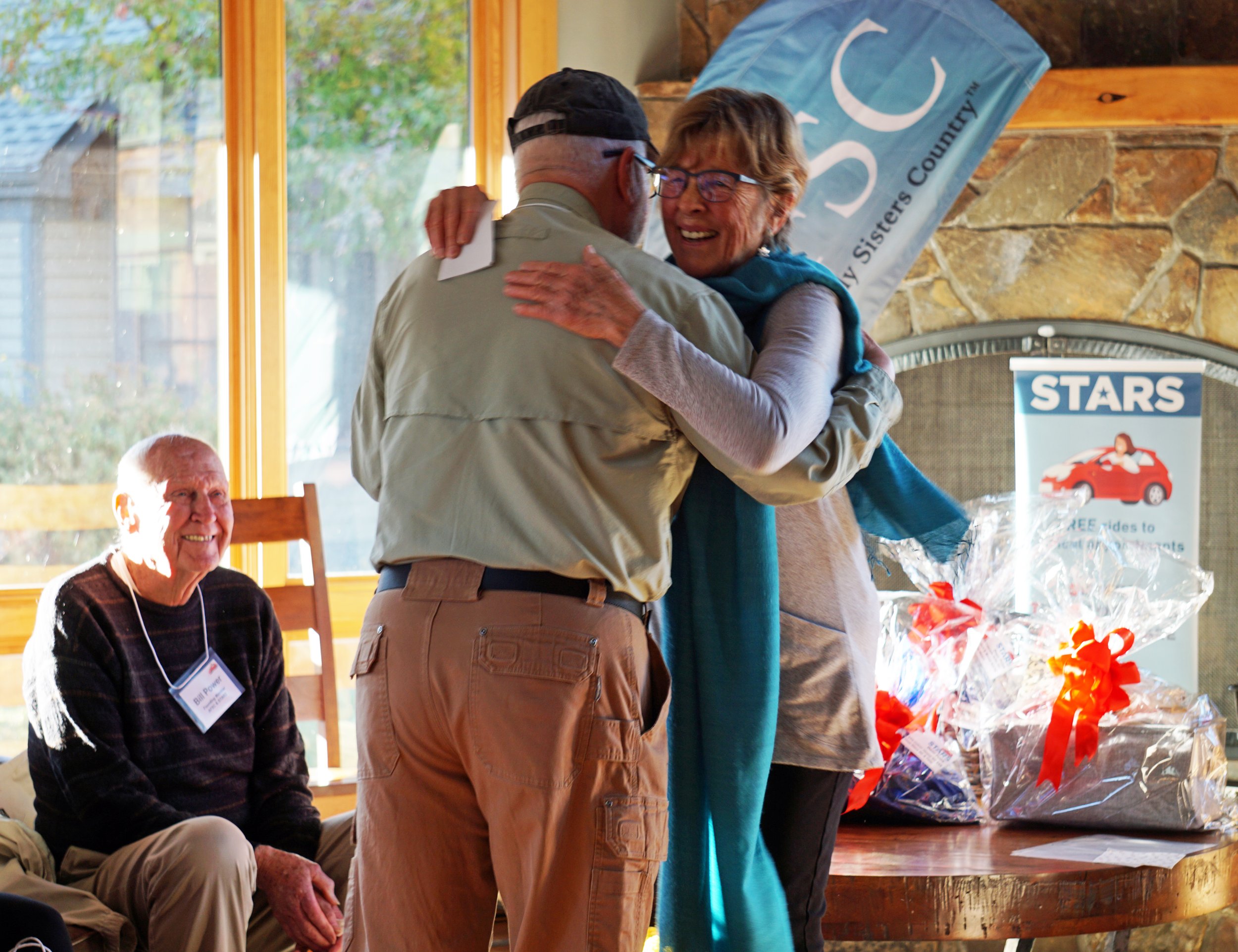 Two people hugging indoors near a window, with a seated person and gift baskets in the background.
