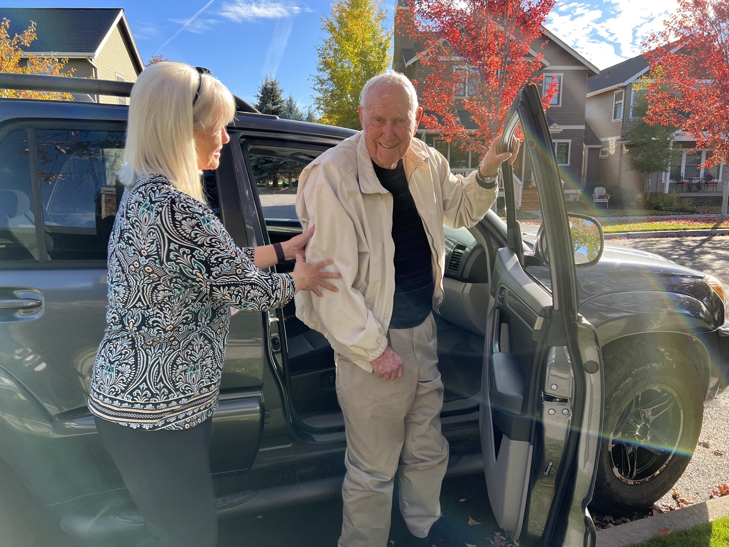 A woman helps an elderly man out of a vehicle on a sunny day with trees and houses in the background.