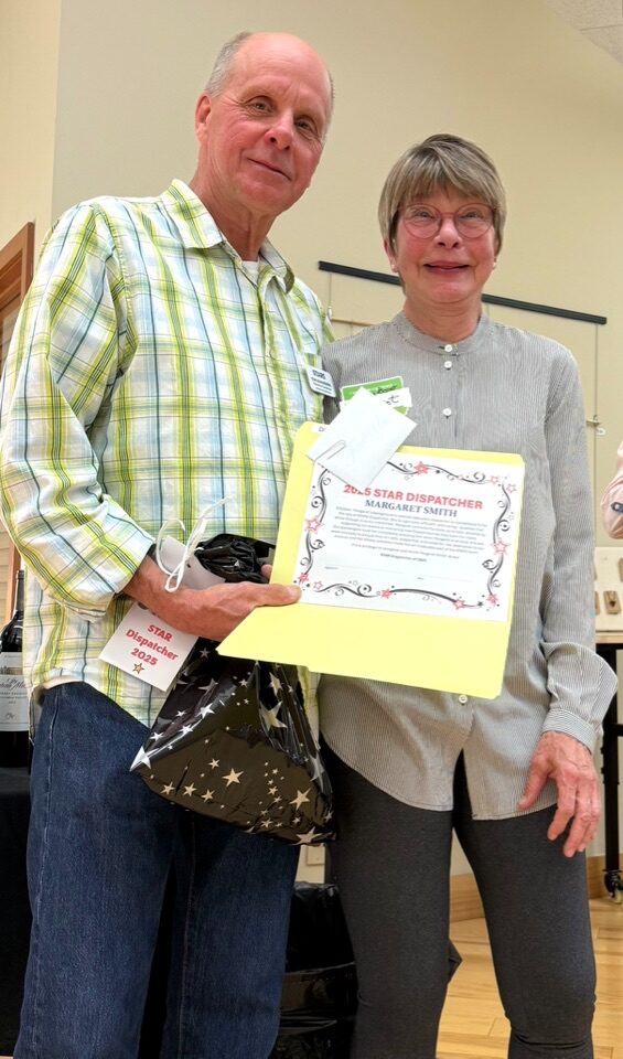 Man and woman holding a certificate and gift bag, smiling indoors, light-colored wall background.