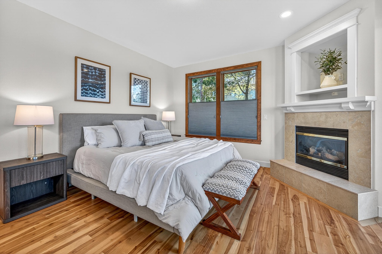 Bedroom with bed, two nightstands, window, fireplace, and artwork, featuring hardwood floors and neutral decor.