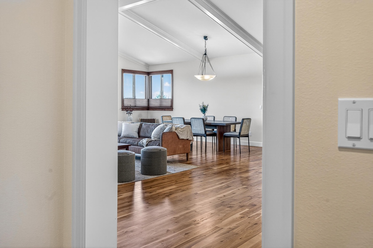 Living room with wooden floor, sofa, chairs, window, and hanging light fixture, viewed through doorway.