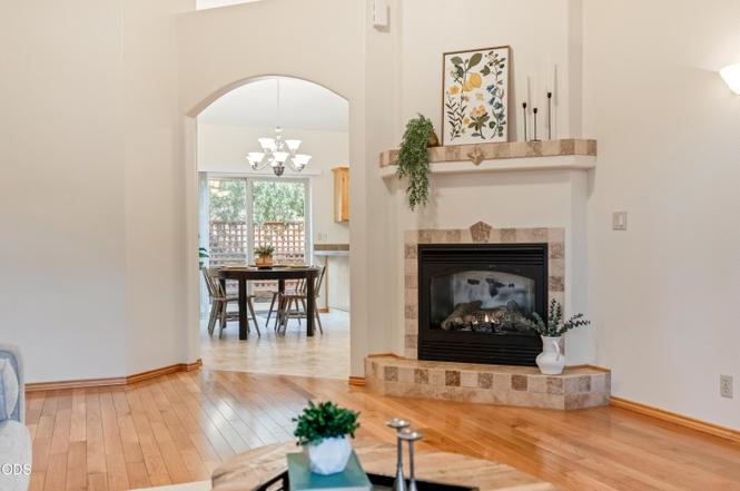 Living room with fireplace, mantel decorated with plants and artwork, hardwood floor, and view into dining area with table and chairs.
