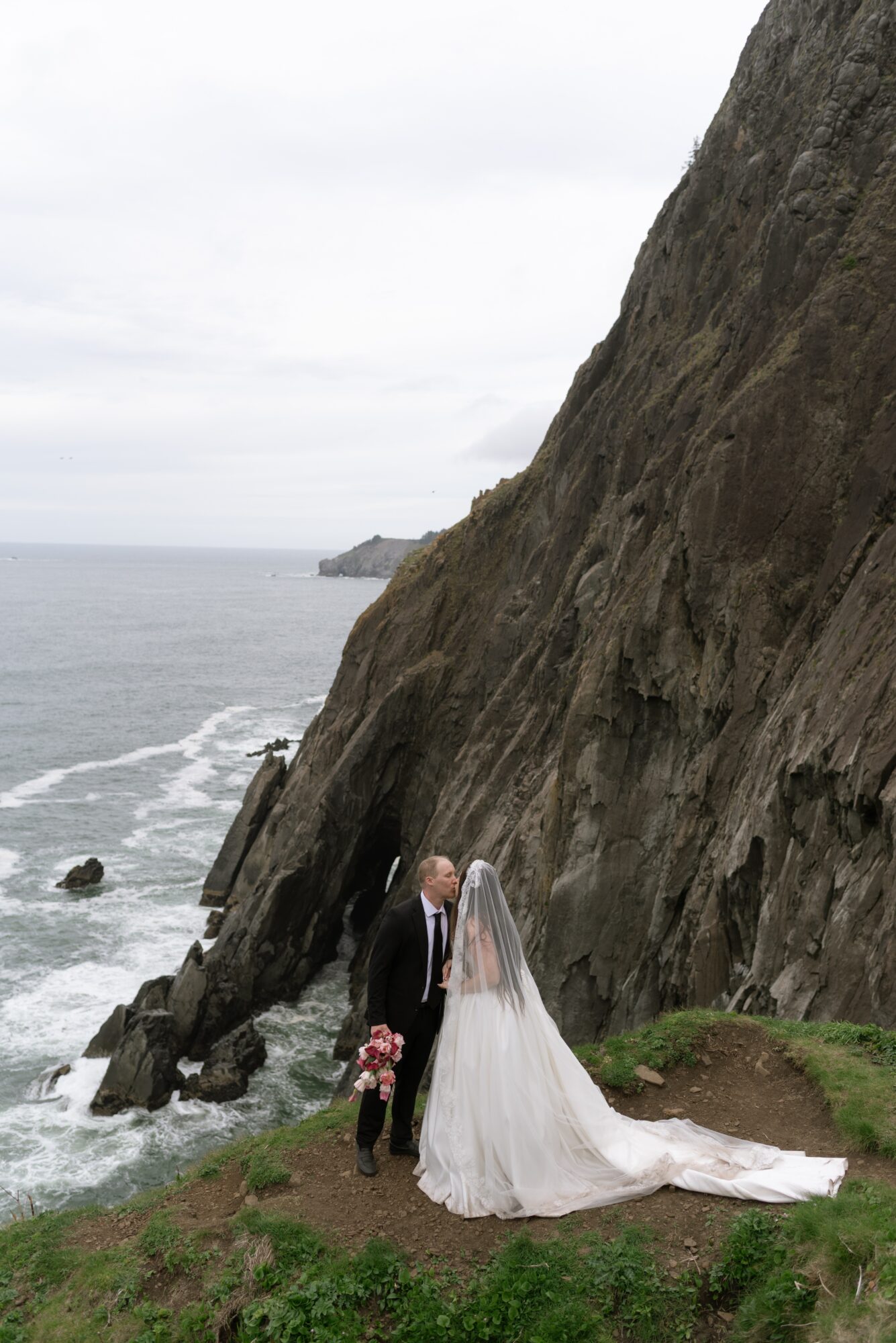 Bride and groom standing near a rocky cliff by the ocean, with a child holding flowers, overcast sky, grassy area.