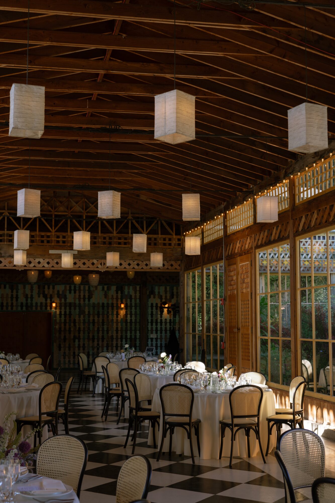 Elegant dining area with round tables, chairs, and hanging lanterns inside a wooden structure with large windows.