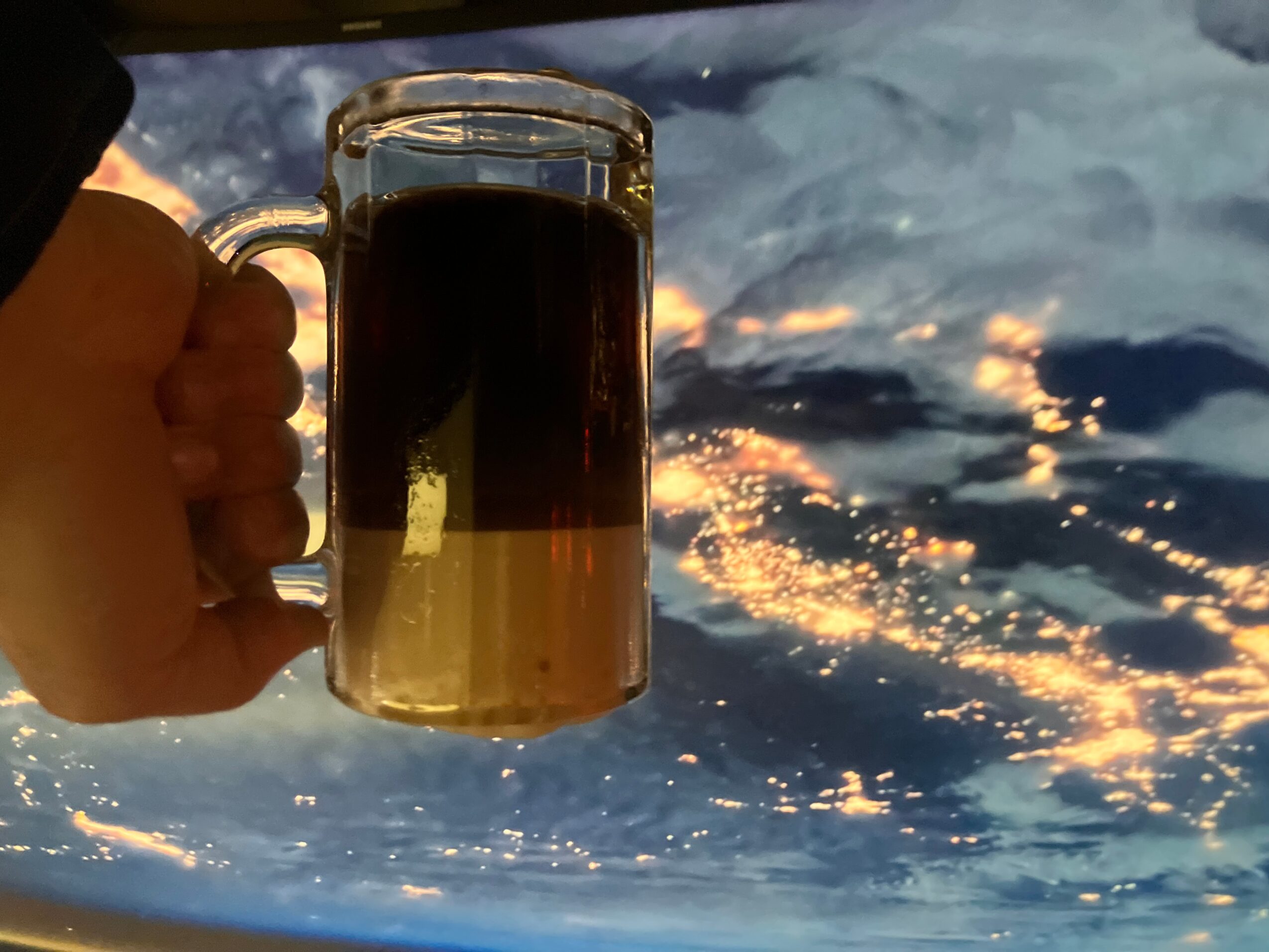 Person holding a glass mug of dark beverage against a cloudy sky with sunlight reflections.