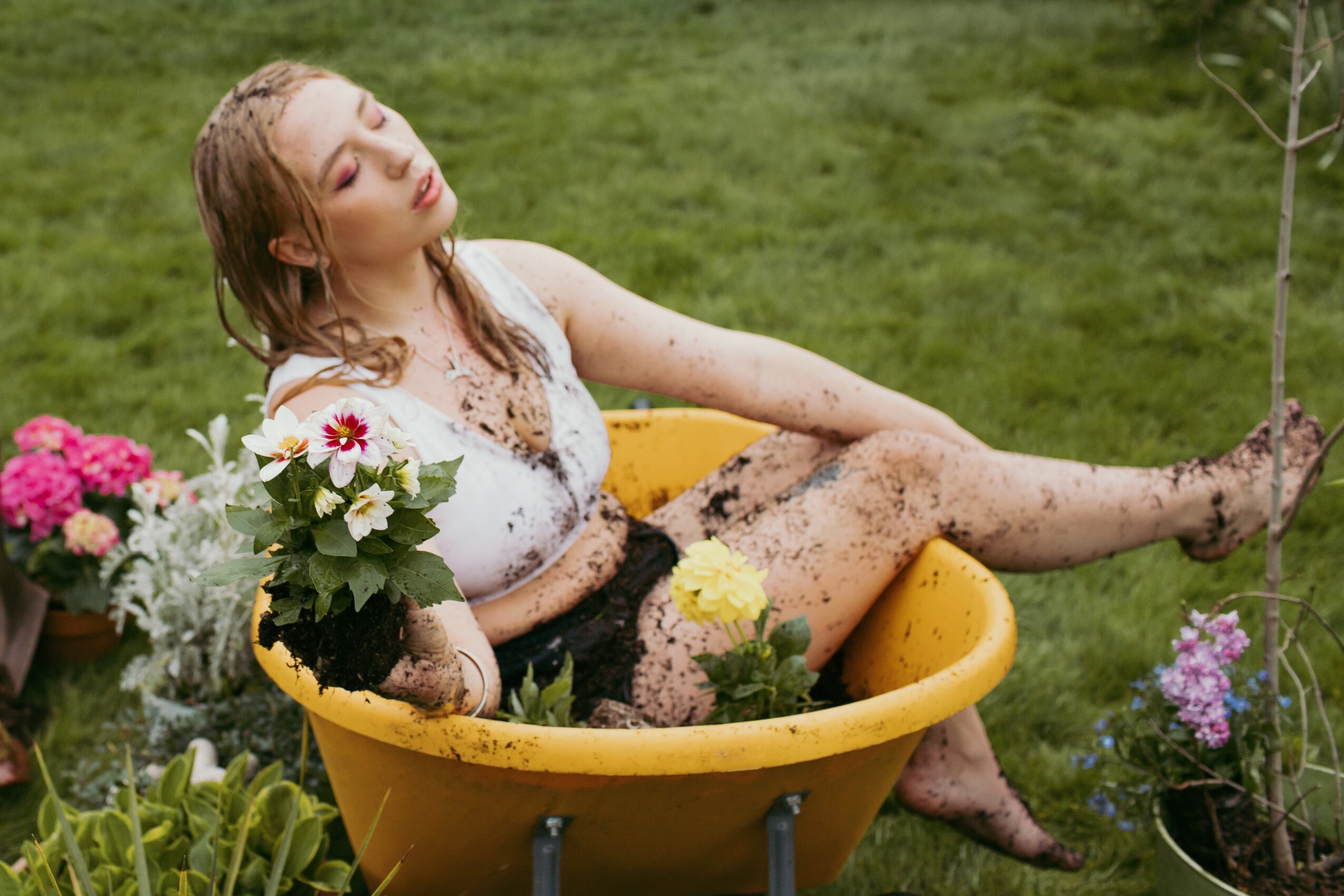 Woman sitting in a yellow bathtub outdoors, surrounded by flowers and greenery, with mud on her legs and arms.