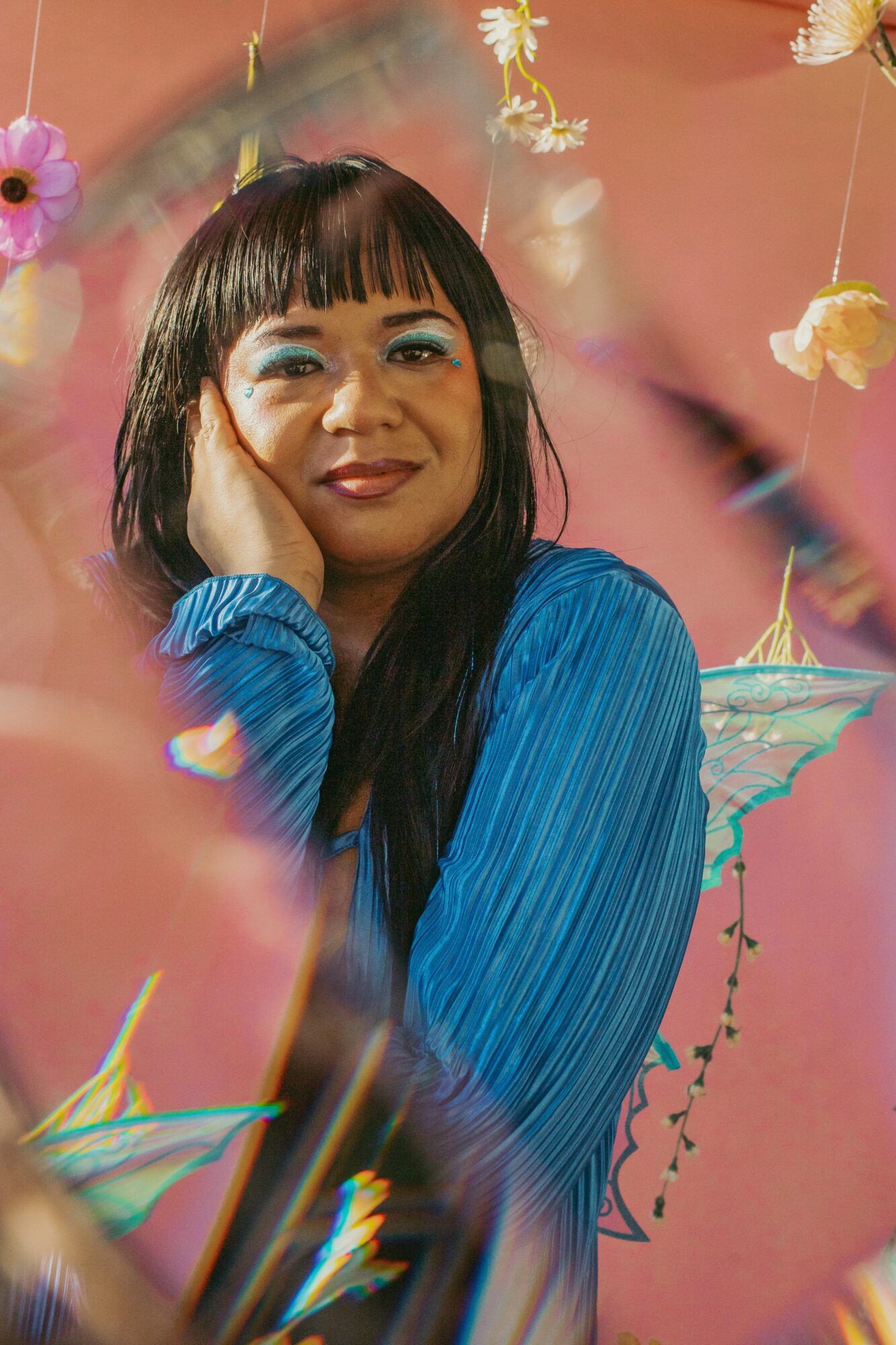 Woman with long dark hair and blue makeup, wearing a blue top, surrounded by hanging flowers and pink background.