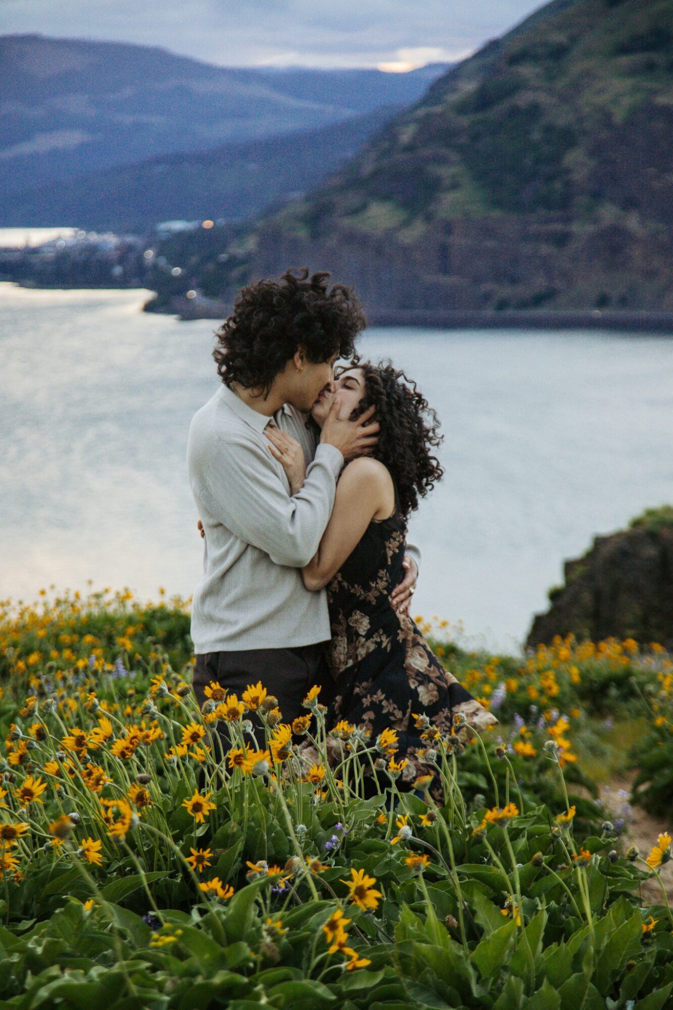A couple embraces in a field of yellow flowers near a body of water with mountains in the background.