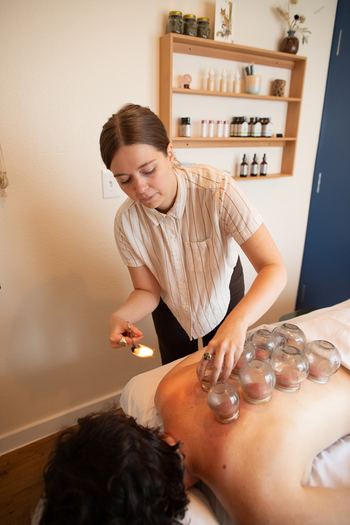 Woman performing a cupping therapy on a person's back in a room with shelves of bottles and jars.