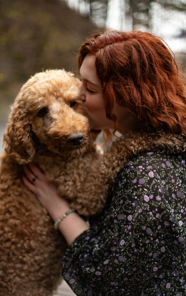Woman with red hair holding a curly-haired dog outdoors, close-up view, natural background, affectionate interaction.