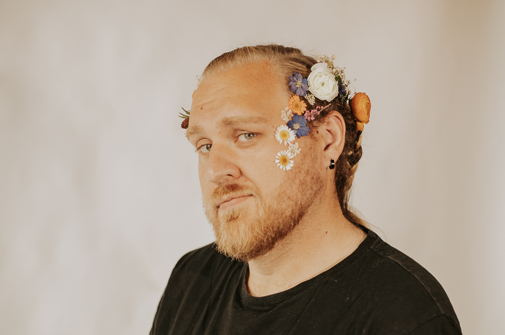 Man with a beard and long hair decorated with colorful flowers and small objects, looking at the camera.