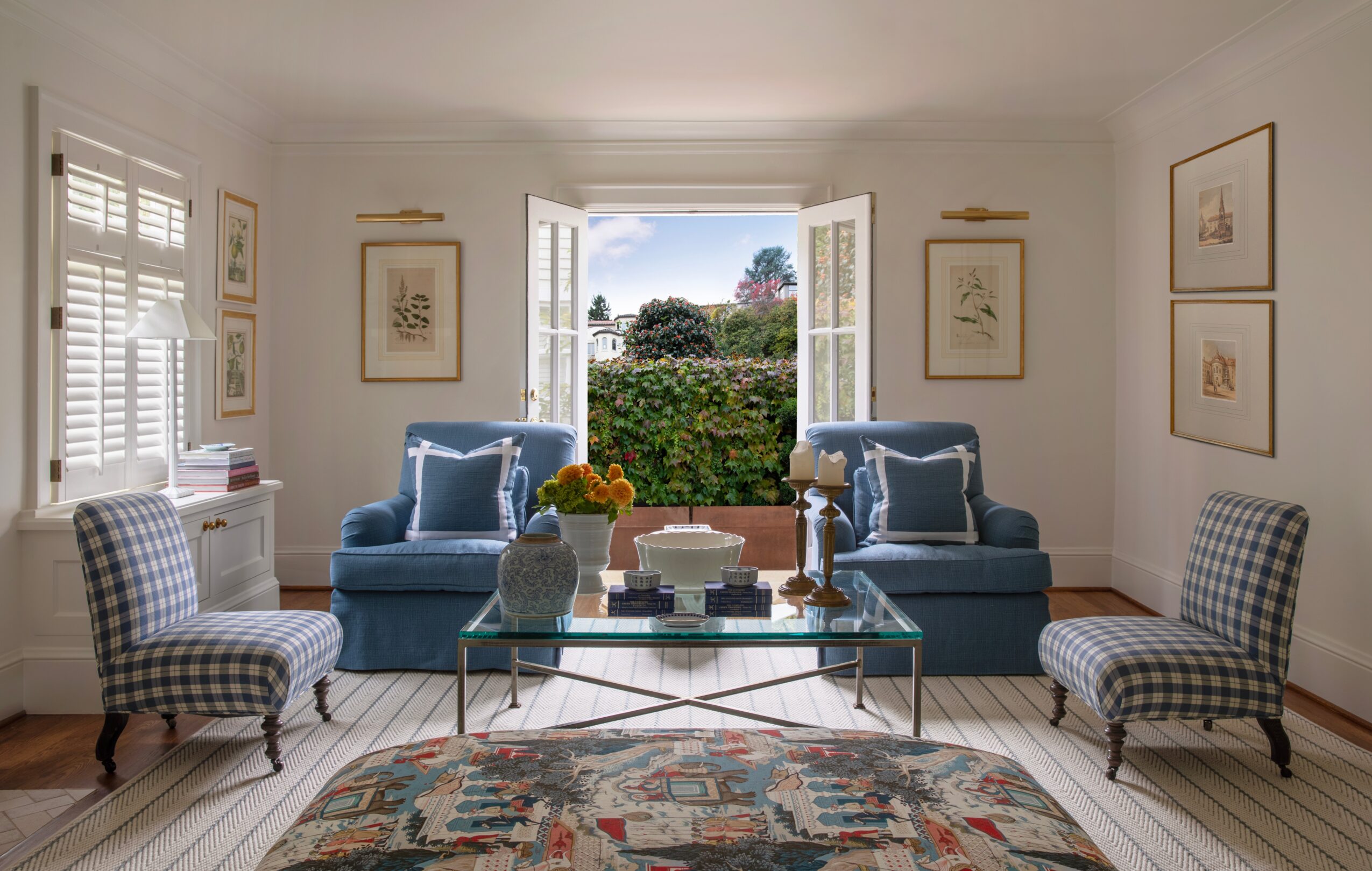 Living room with two blue sofas, checkered armchairs, glass coffee table, and a large window showing greenery outside.