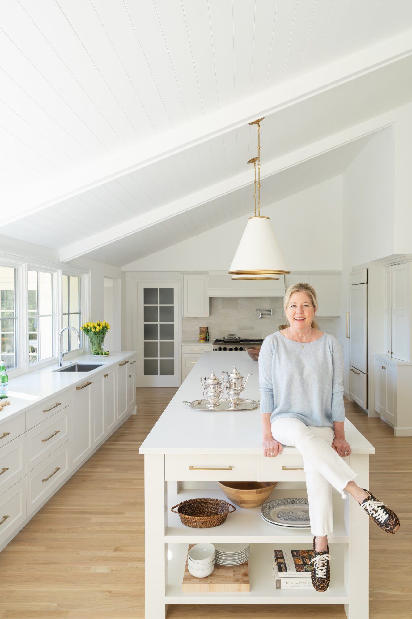 Woman sitting on kitchen island in bright, modern kitchen with white cabinets and large windows, smiling, with a vase of flowers nearby.