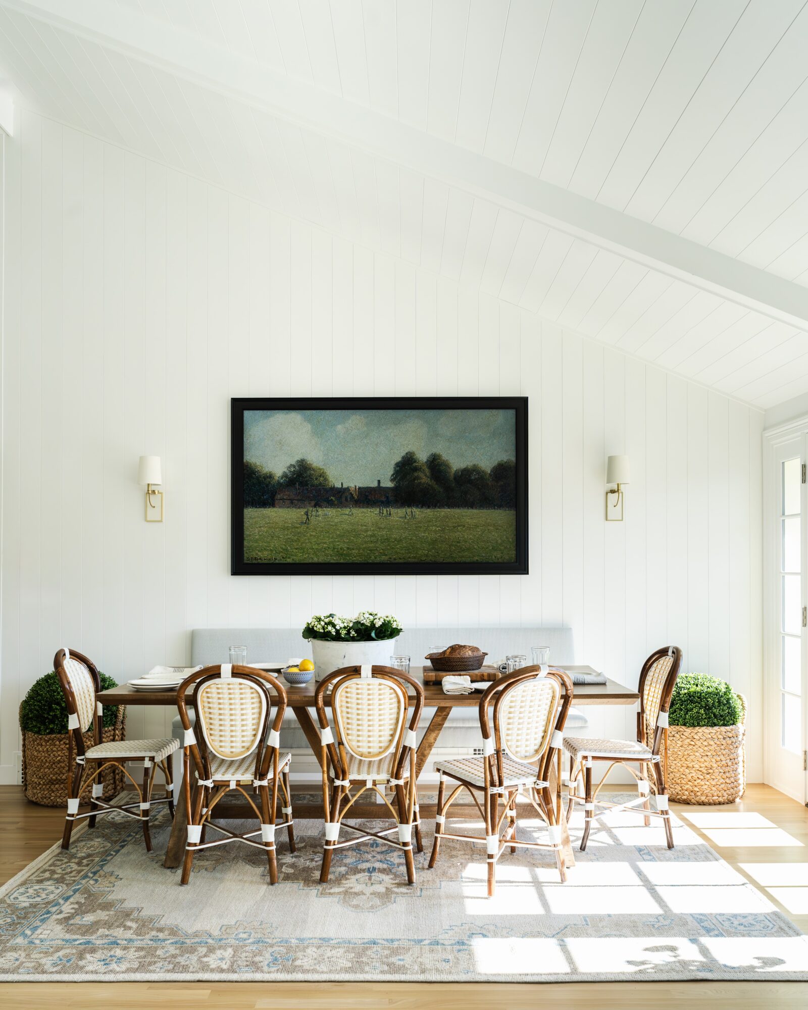 Dining room with a large table, eight chairs, a framed landscape painting, and sunlight streaming through windows.