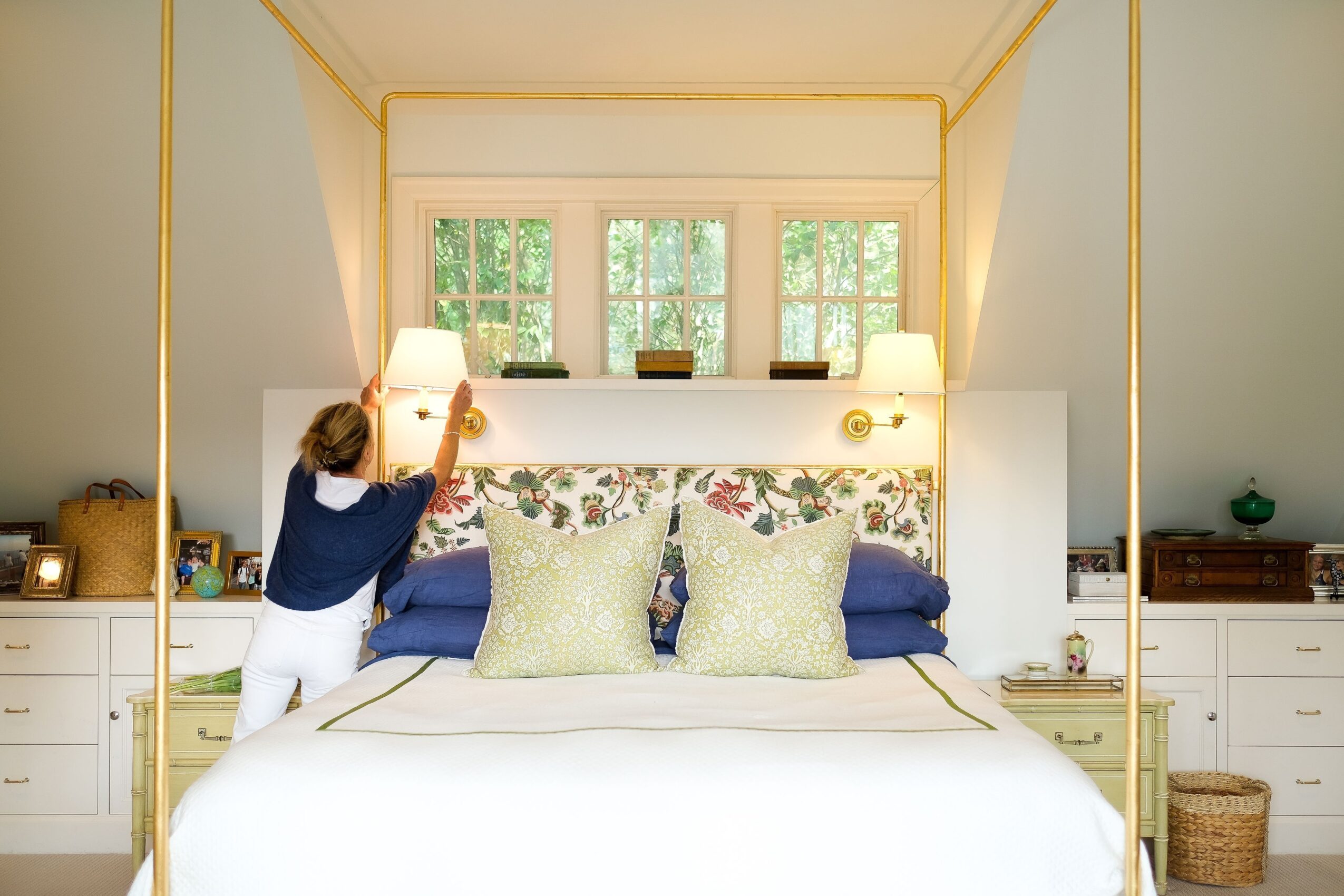 Person adjusting a lamp on a bed with pillows in a bright bedroom with large windows, white walls, and side tables.