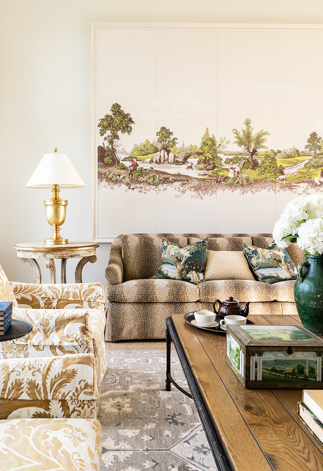 Living room with a beige sofa, patterned armchair, side table with lamp, large landscape painting, and wooden coffee table with decorative items.