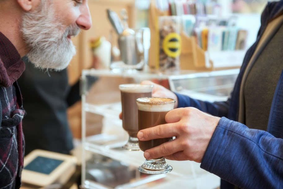 Two people holding coffee drinks in a cafe, with a blurred background of shelves and a counter.
