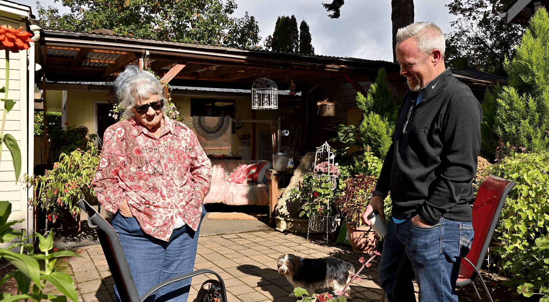 An elderly woman and a man stand outdoors in a garden with plants and furniture, talking and smiling, with a dog nearby.