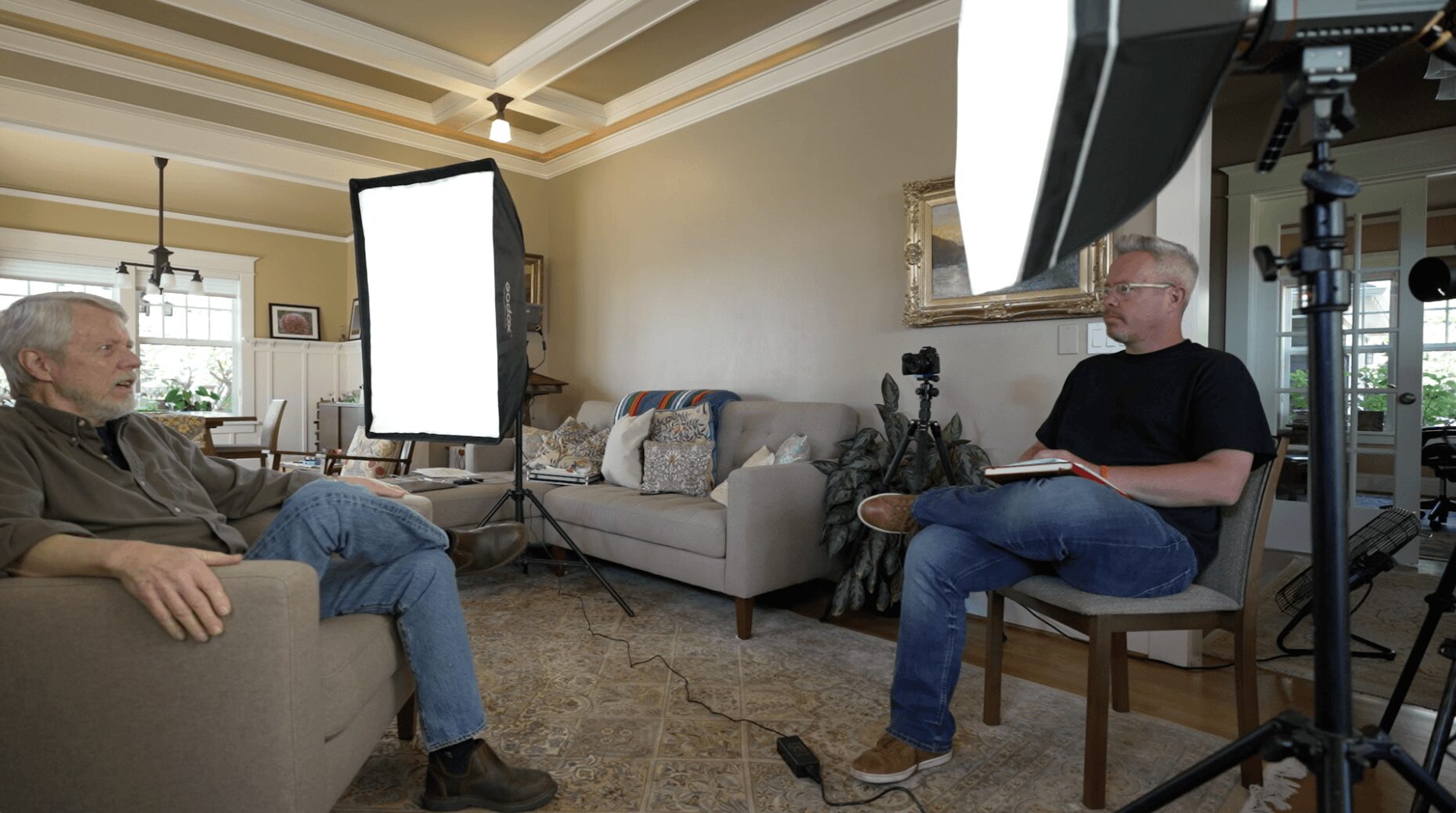 Two men sit in a living room with lighting equipment and a camera, engaged in conversation, with a sofa and window in background.