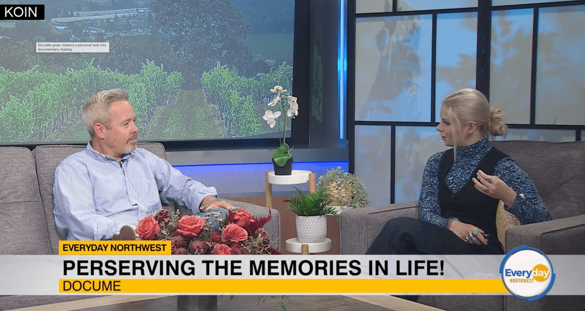 Two people sit on a talk show set with plants and a large window behind them, discussing memories.