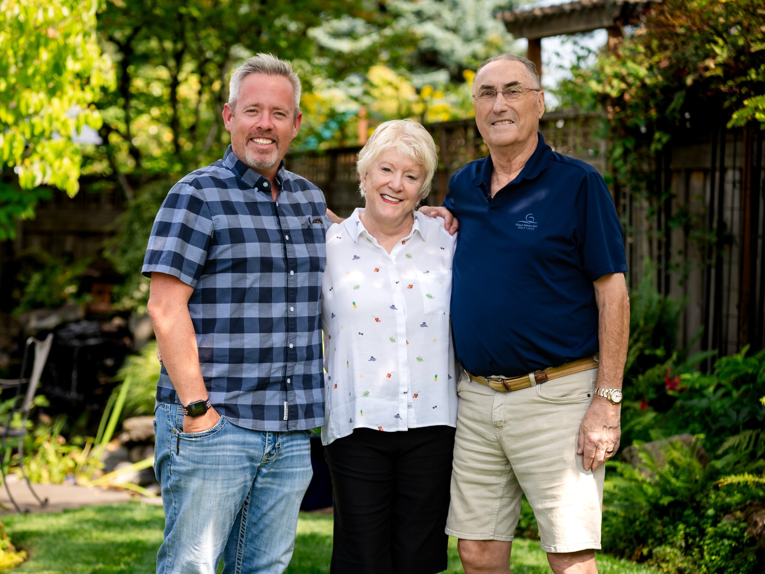 Three people standing outdoors in a garden, smiling, with trees and plants in the background.