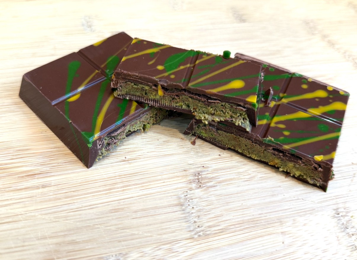 Chocolate bar with green and yellow drizzle on top, partially cut to show green filling inside, on a light wooden surface.