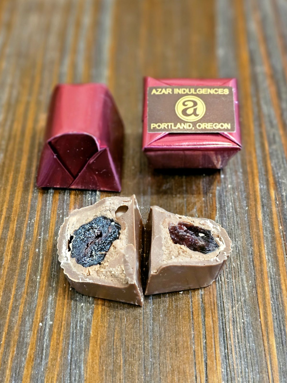 Unwrapped chocolate with dark filling inside, next to a pink box labeled 'Azar Indulgence' on a wooden surface.