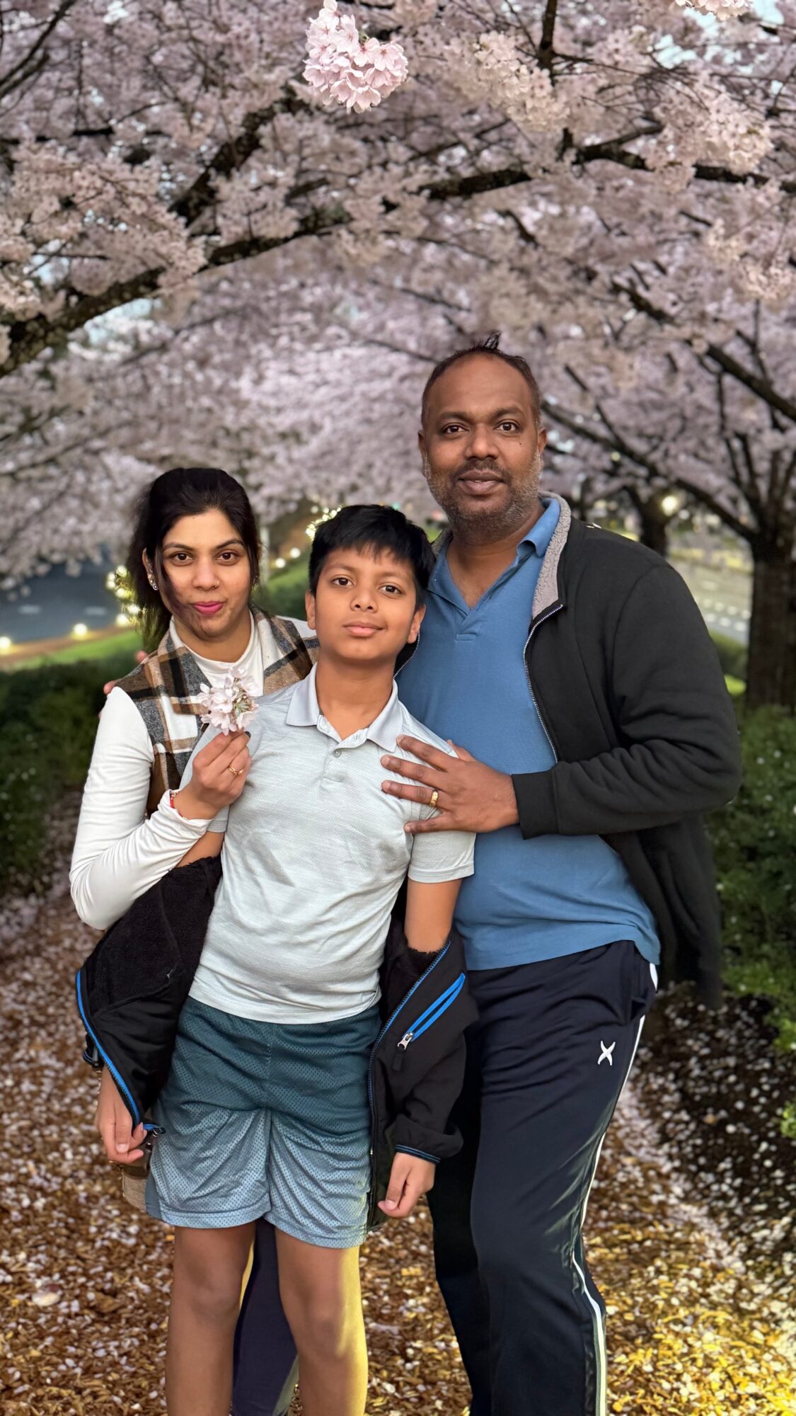 Three people standing outdoors under cherry blossom trees, smiling, with pink flowers in the background.