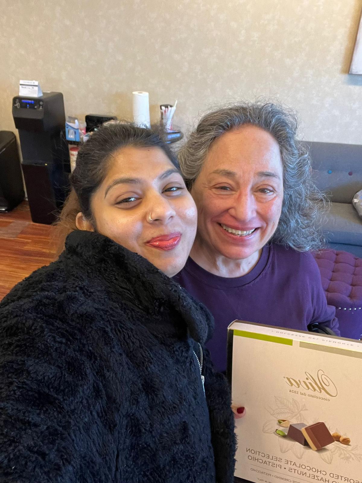 Two women smiling indoors, one with dark hair and the other with gray hair, holding a box of chocolates.