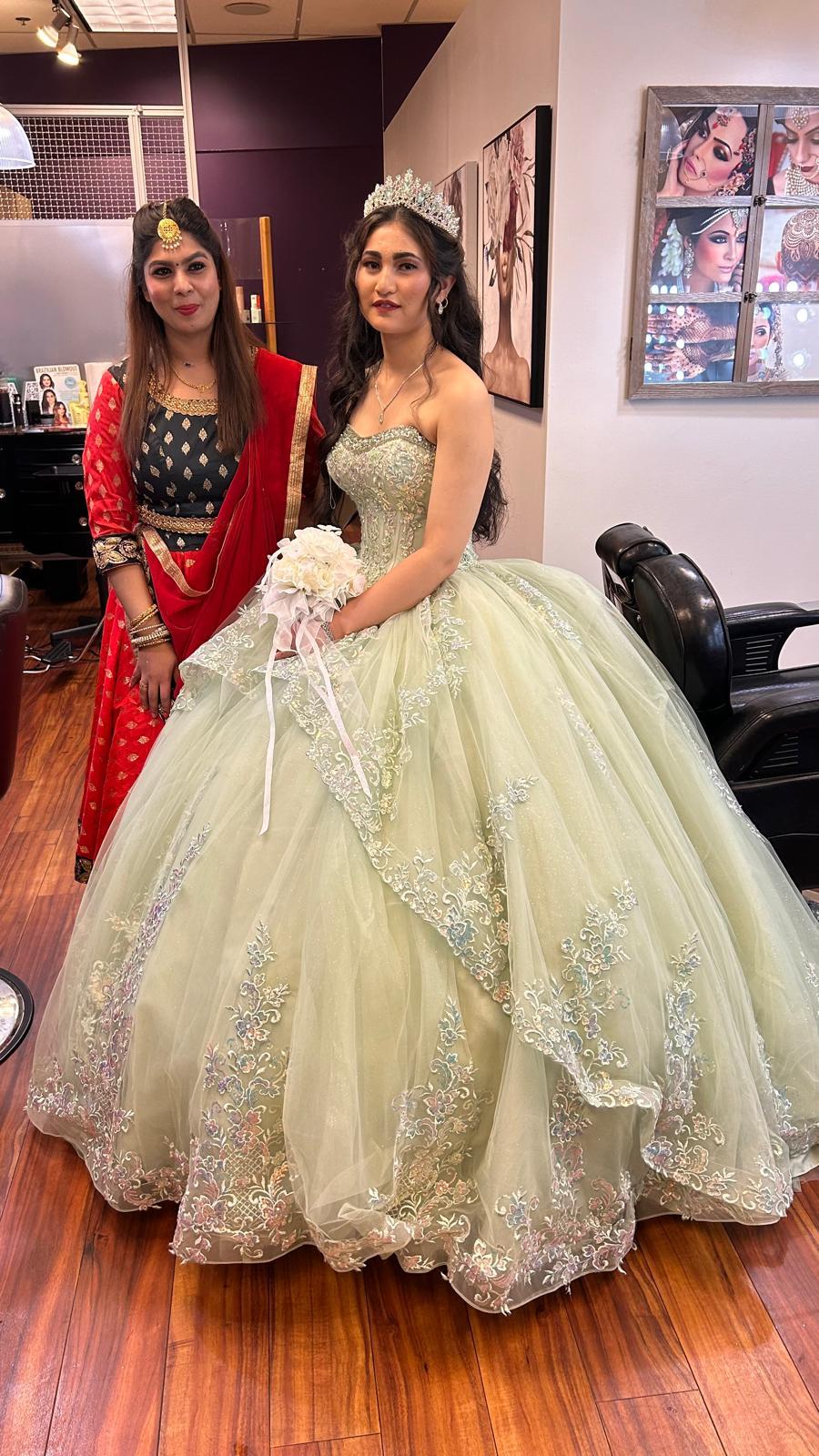 Two women stand indoors, one in a red dress and the other in a light-colored gown with a tiara, in front of framed pictures.
