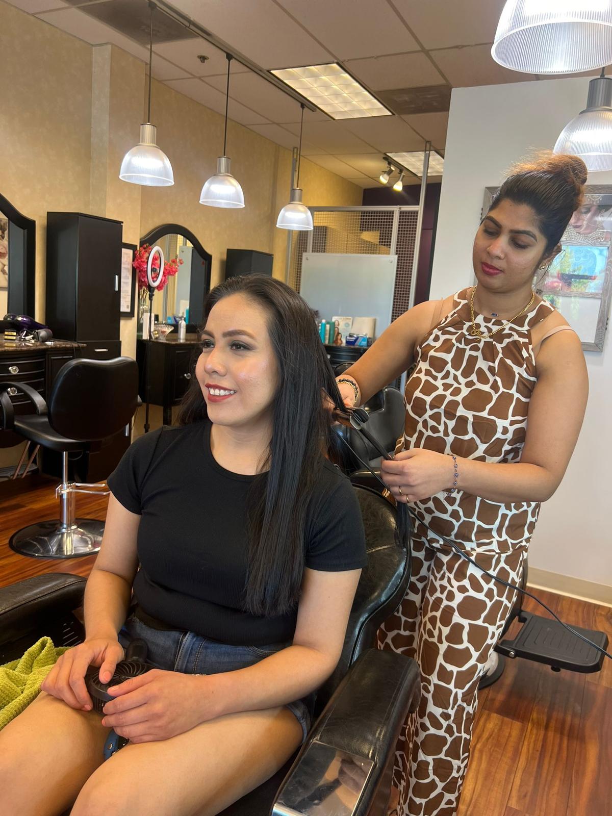 A woman with long dark hair is seated in a salon chair, smiling, while a stylist with a bun styles her hair in a leopard print outfit.