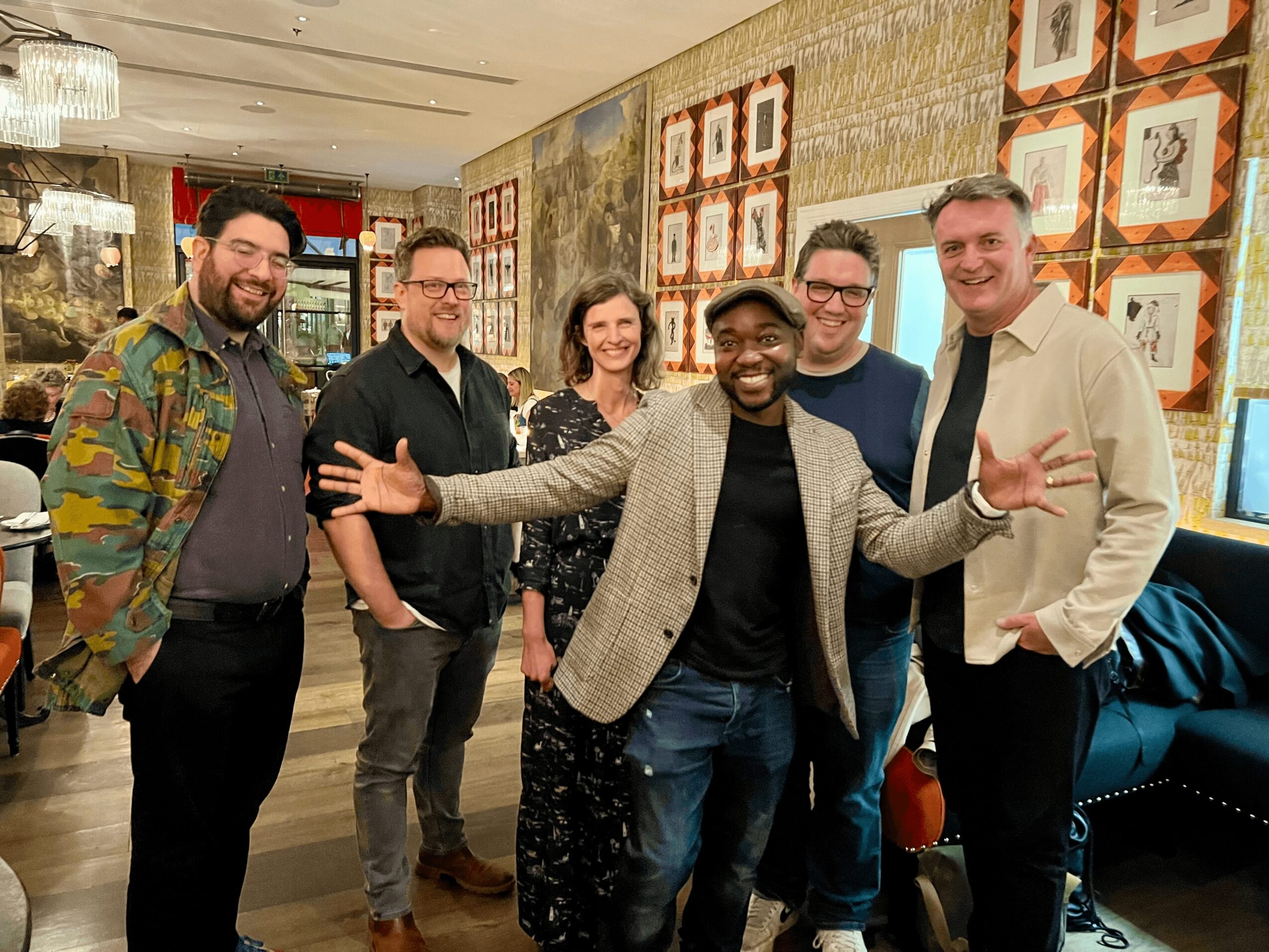 Group of six people smiling and posing together in a decorated room, some with arms around each other.