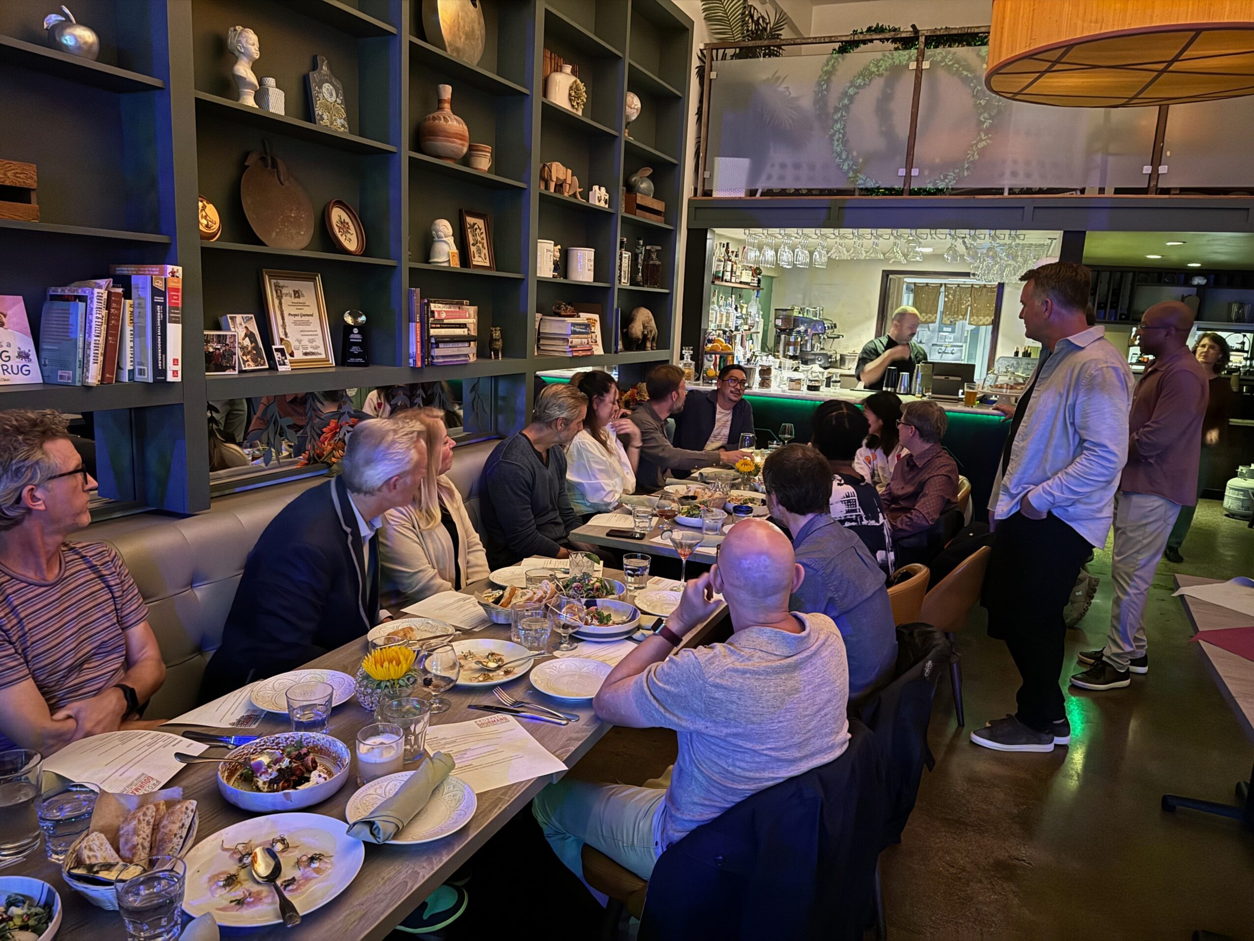 People seated at a long table in a restaurant, with shelves of decorative items and books behind them.
