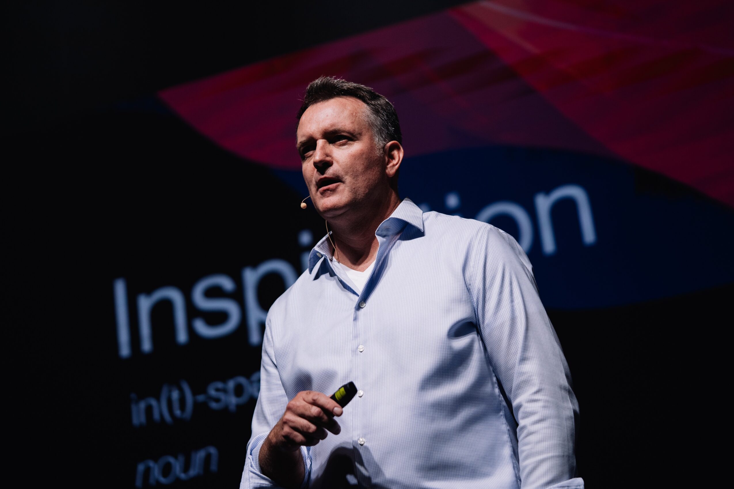 Man in white shirt speaking on stage with a presentation behind him, holding a remote control, dark background.