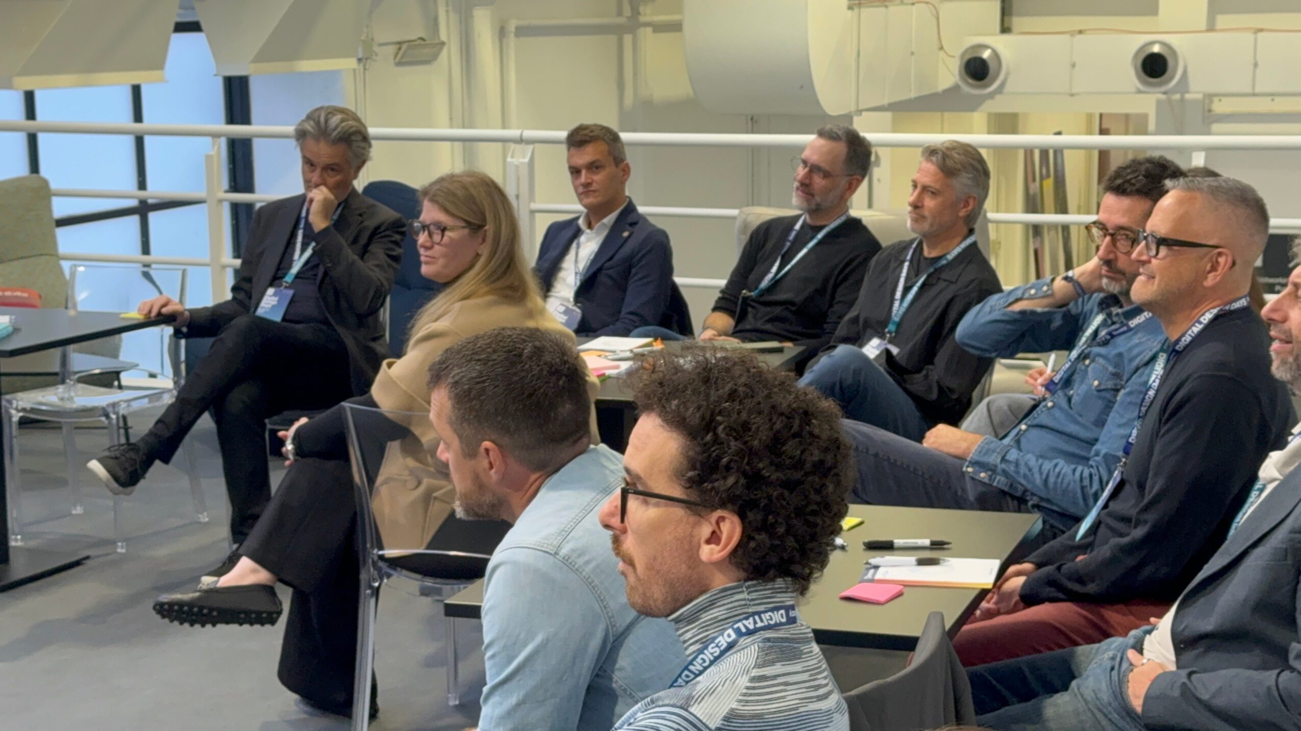 Group of people seated in a modern conference room, listening attentively, with large windows in the background.